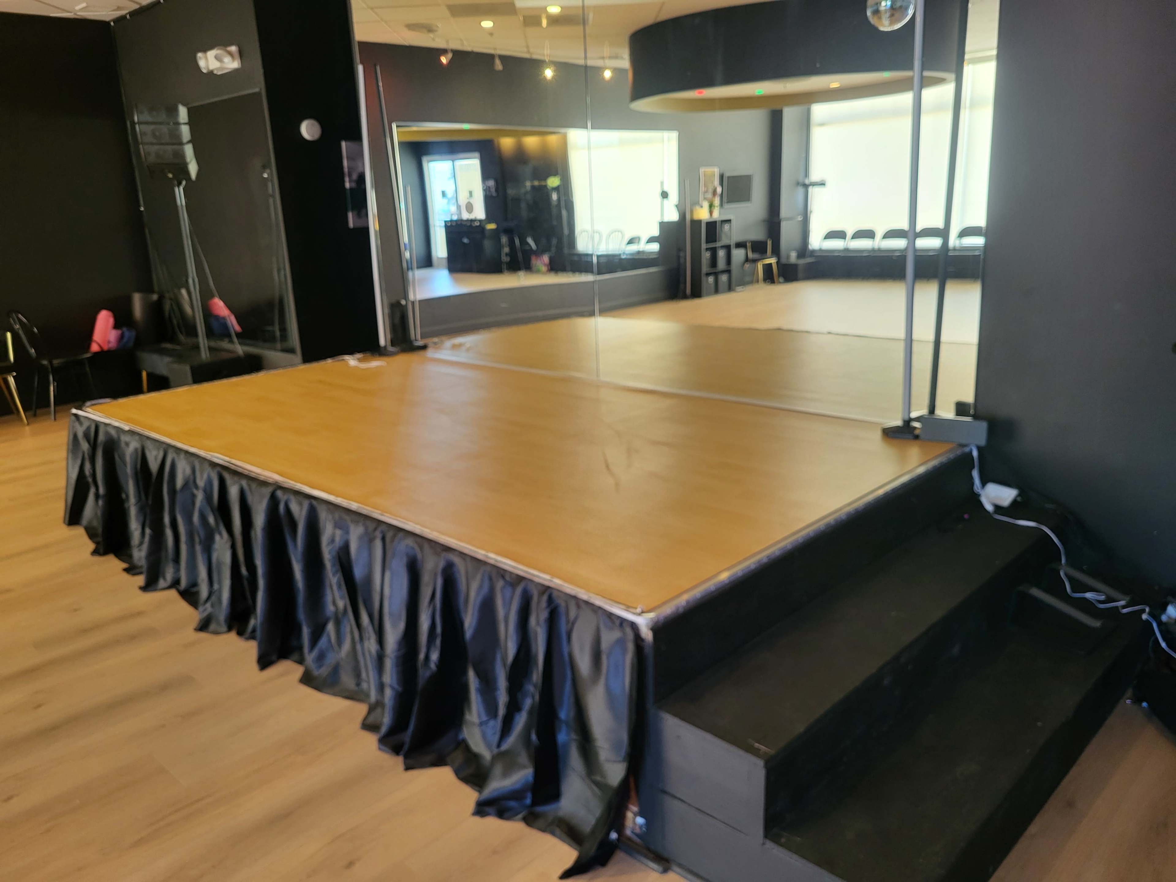 The image shows a wooden stage platform with a black skirt and steps, situated in a dance studio with mirrors and chairs in the background.