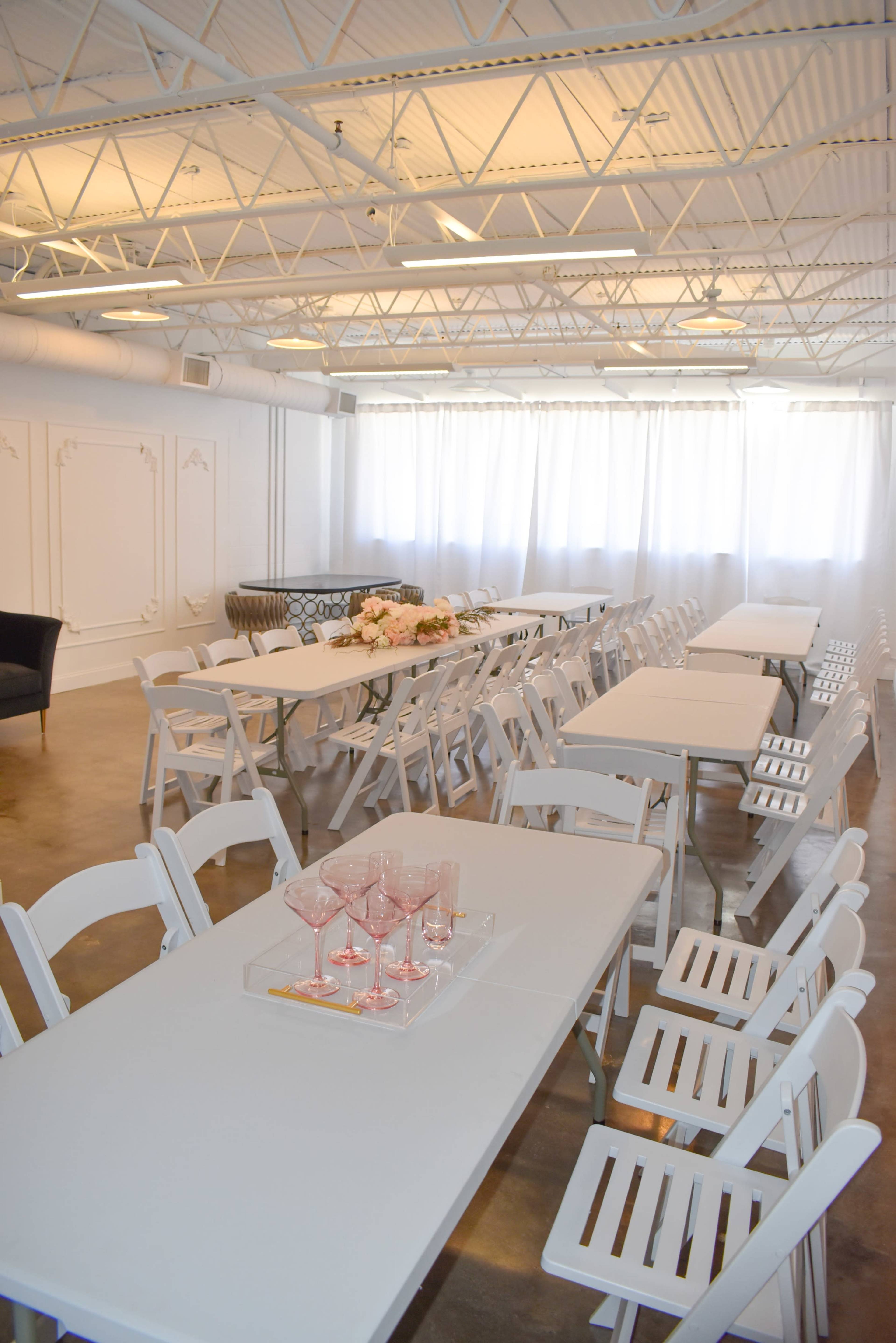 The image shows a spacious, well-lit event room arranged with white tables and chairs, featuring a decorative floral centerpiece and a set of pink glasses on a tray.