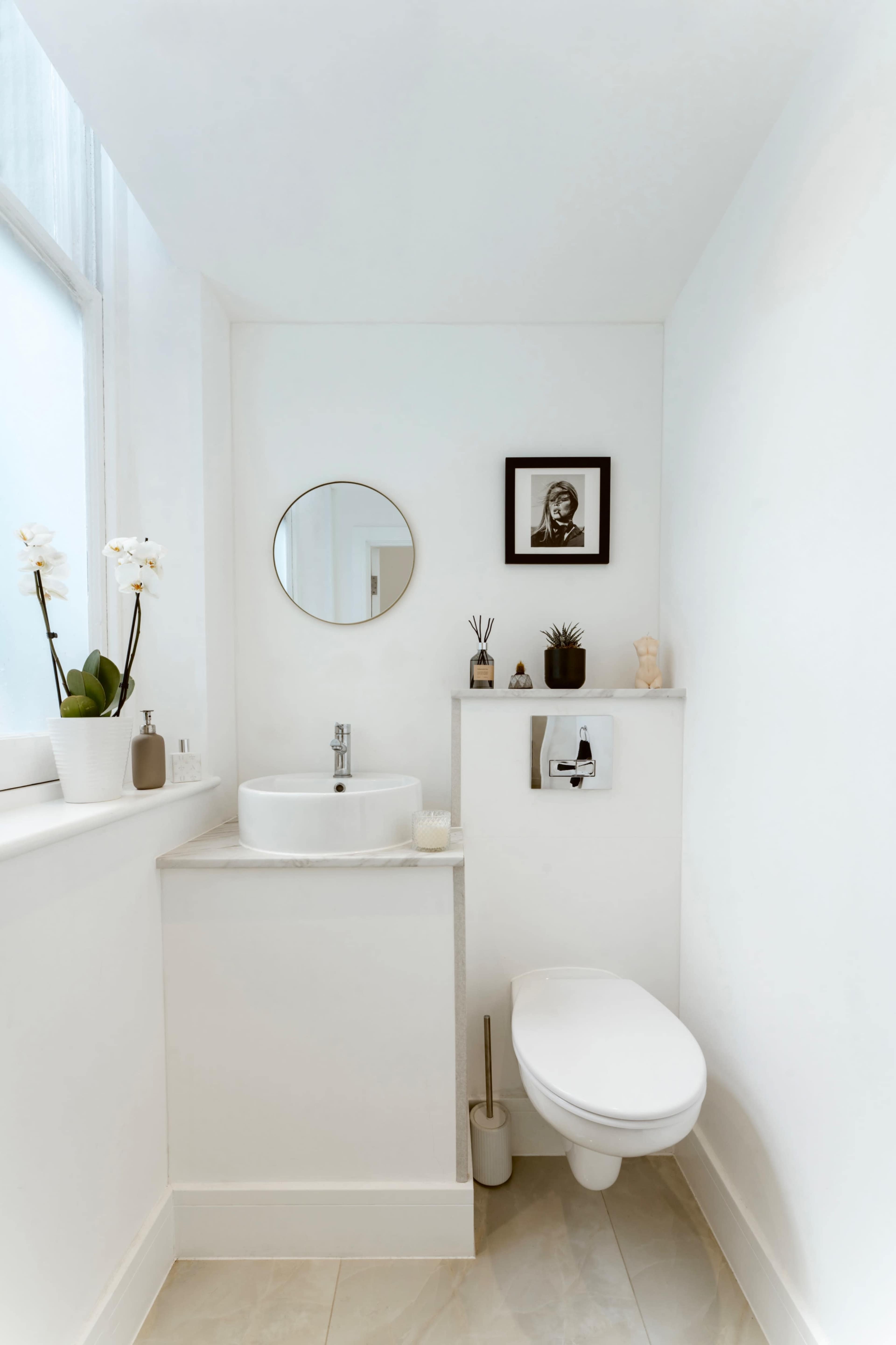 The image shows a small, modern bathroom with a round mirror, a sink, a toilet, and minimal decorative items on the walls and counter.