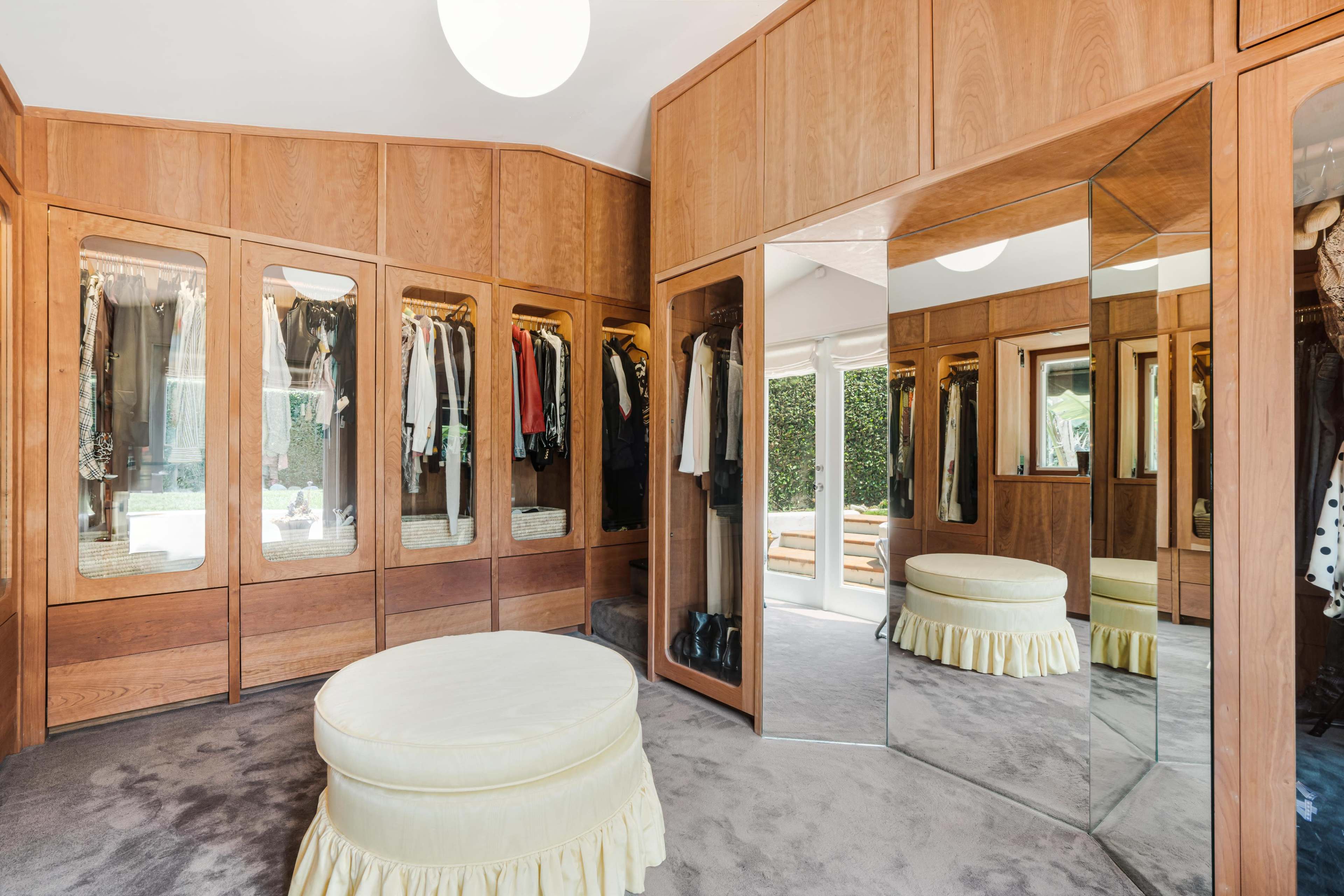 The image shows a walk-in closet featuring wooden cabinetry with clothing displayed and a circular, upholstered ottoman in the center, adjacent to large mirrors and windows.