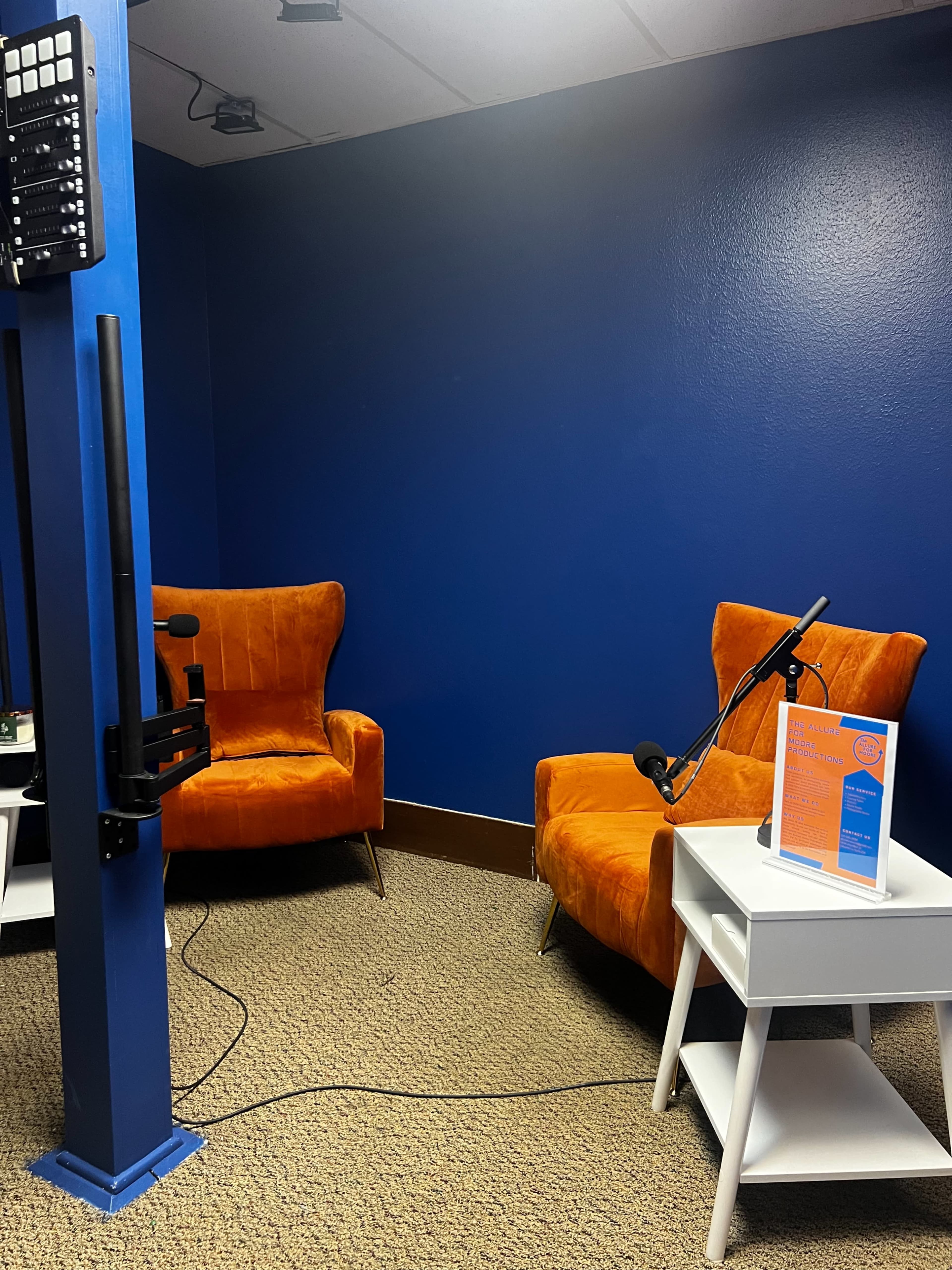 The image shows a small recording studio with two orange chairs, a white side table, and a blue wall.