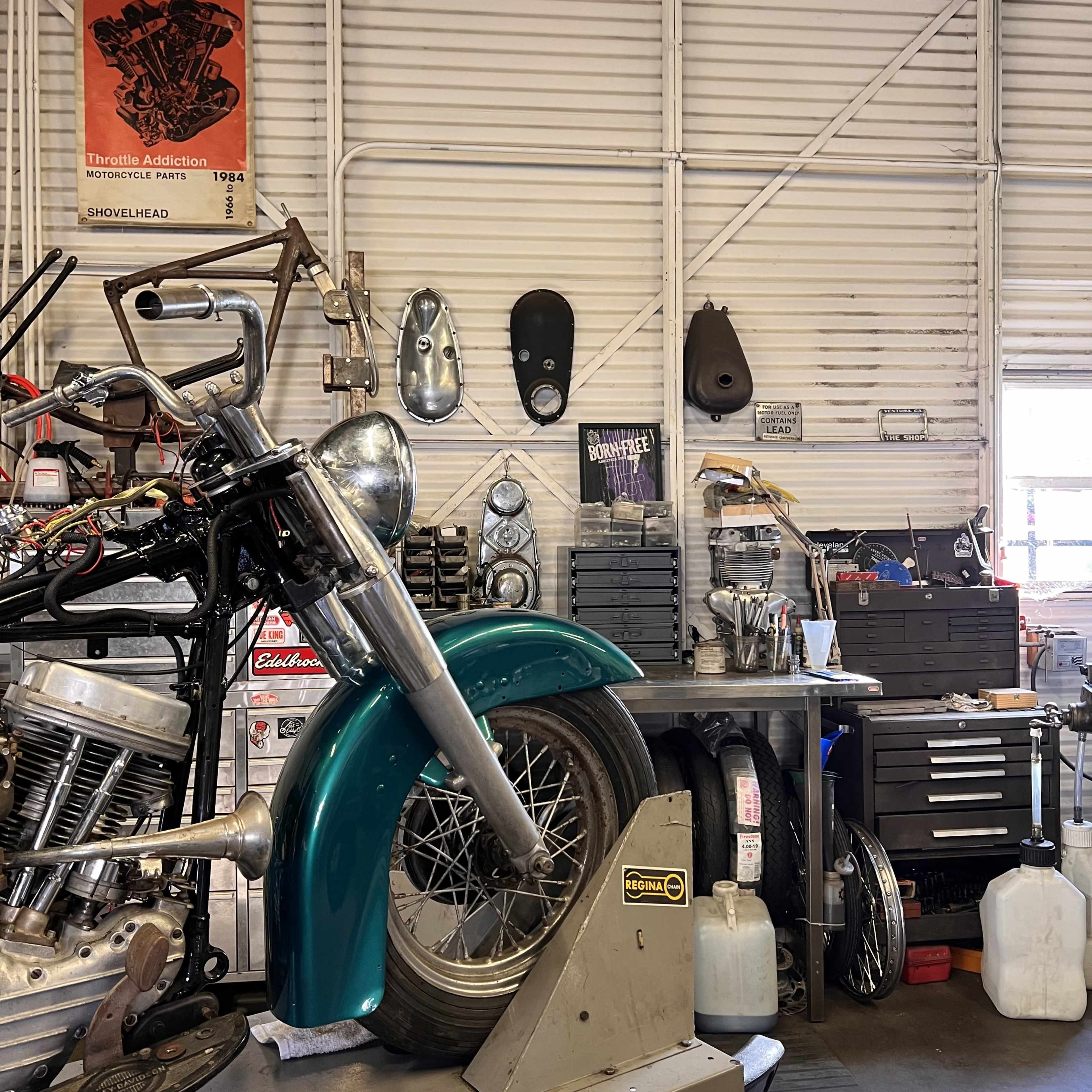 The image shows a motorcycle partially disassembled in a workshop with various tools, parts, and supplies organized on shelves and work surfaces nearby.