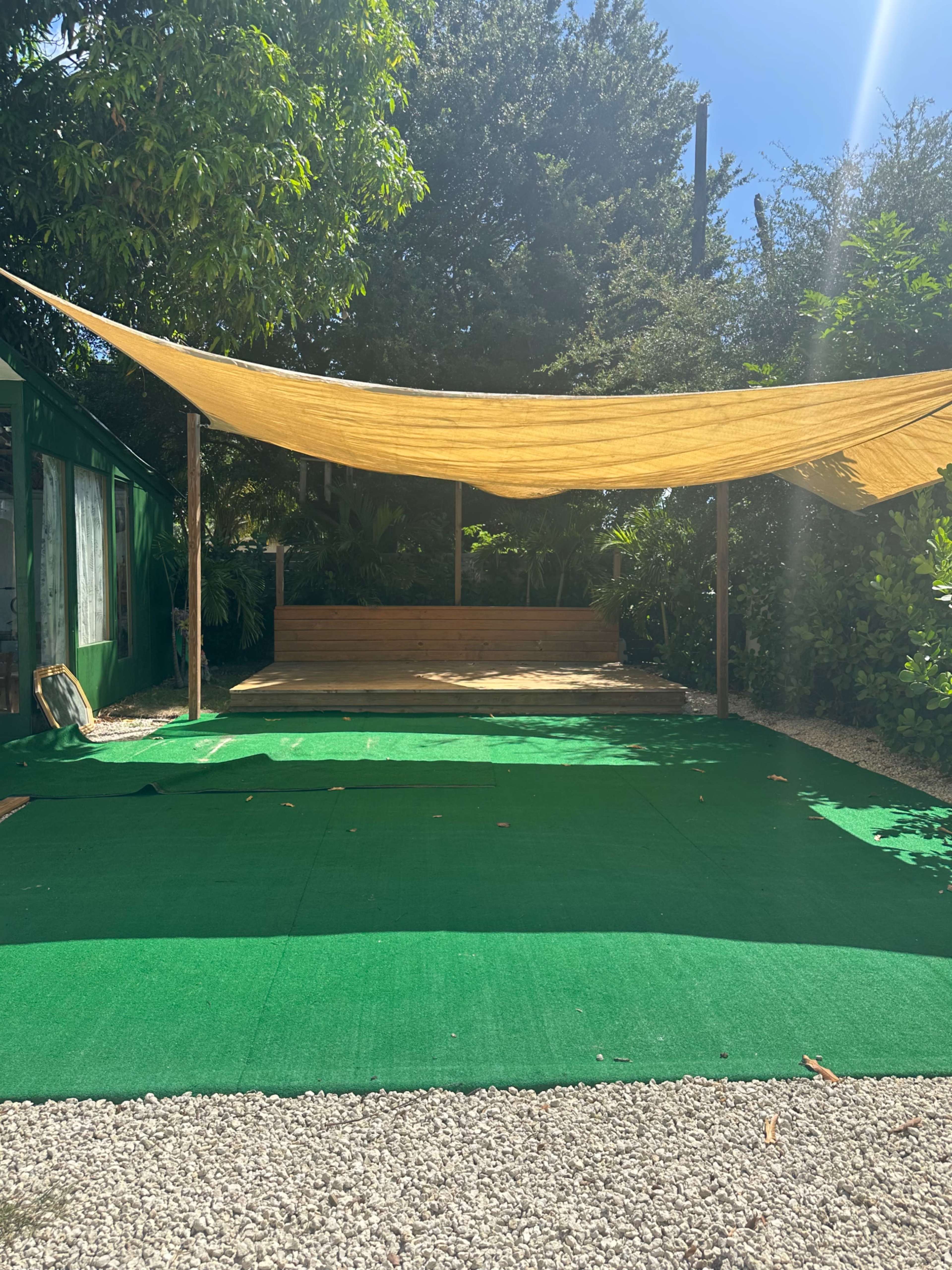 A shaded outdoor area features a wooden bench and green flooring surrounded by trees and foliage.