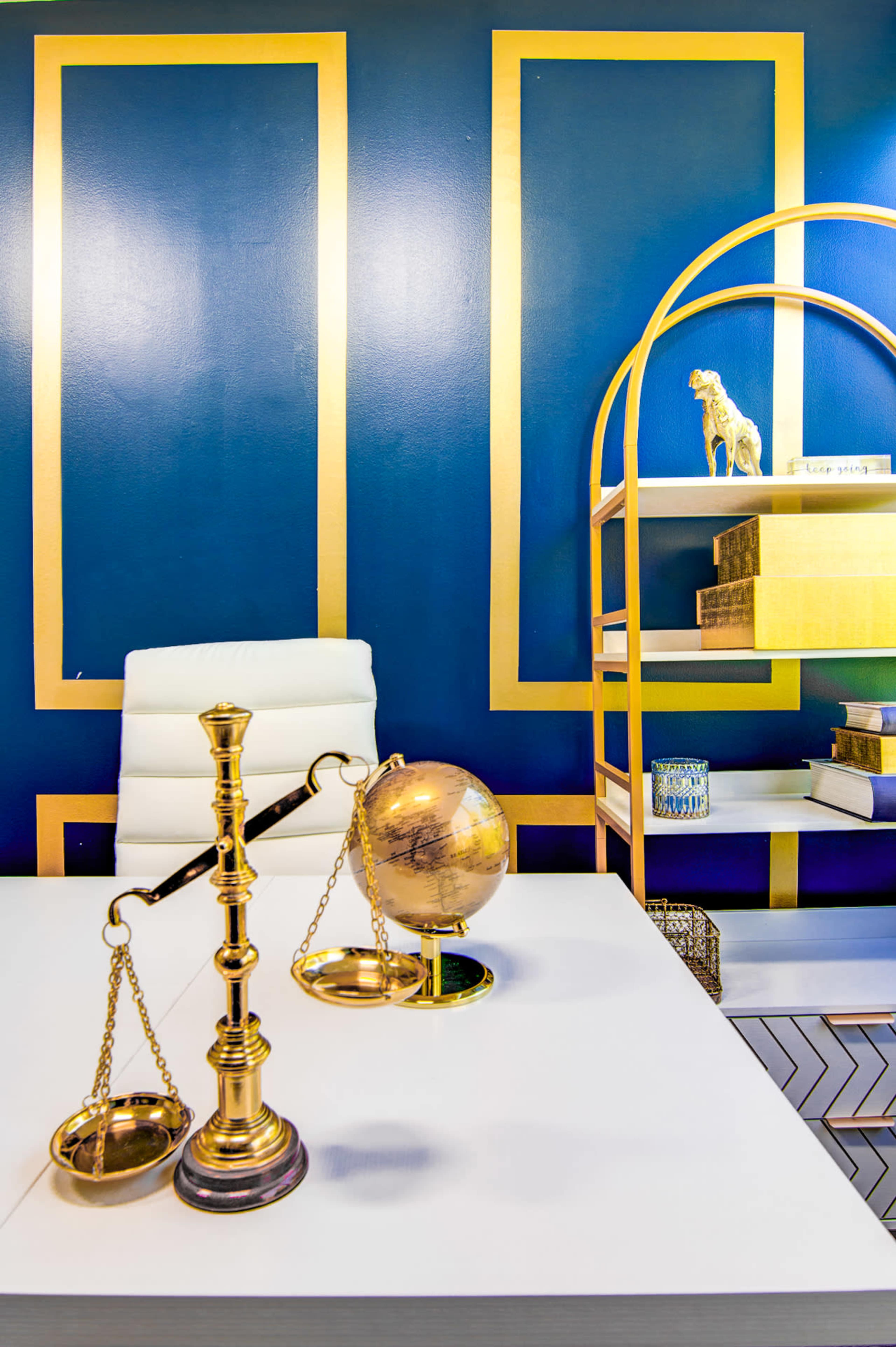 A modern office space with a white desk, a brass balance scale, a globe, and a golden bookshelf against a blue wall featuring gold geometric designs.