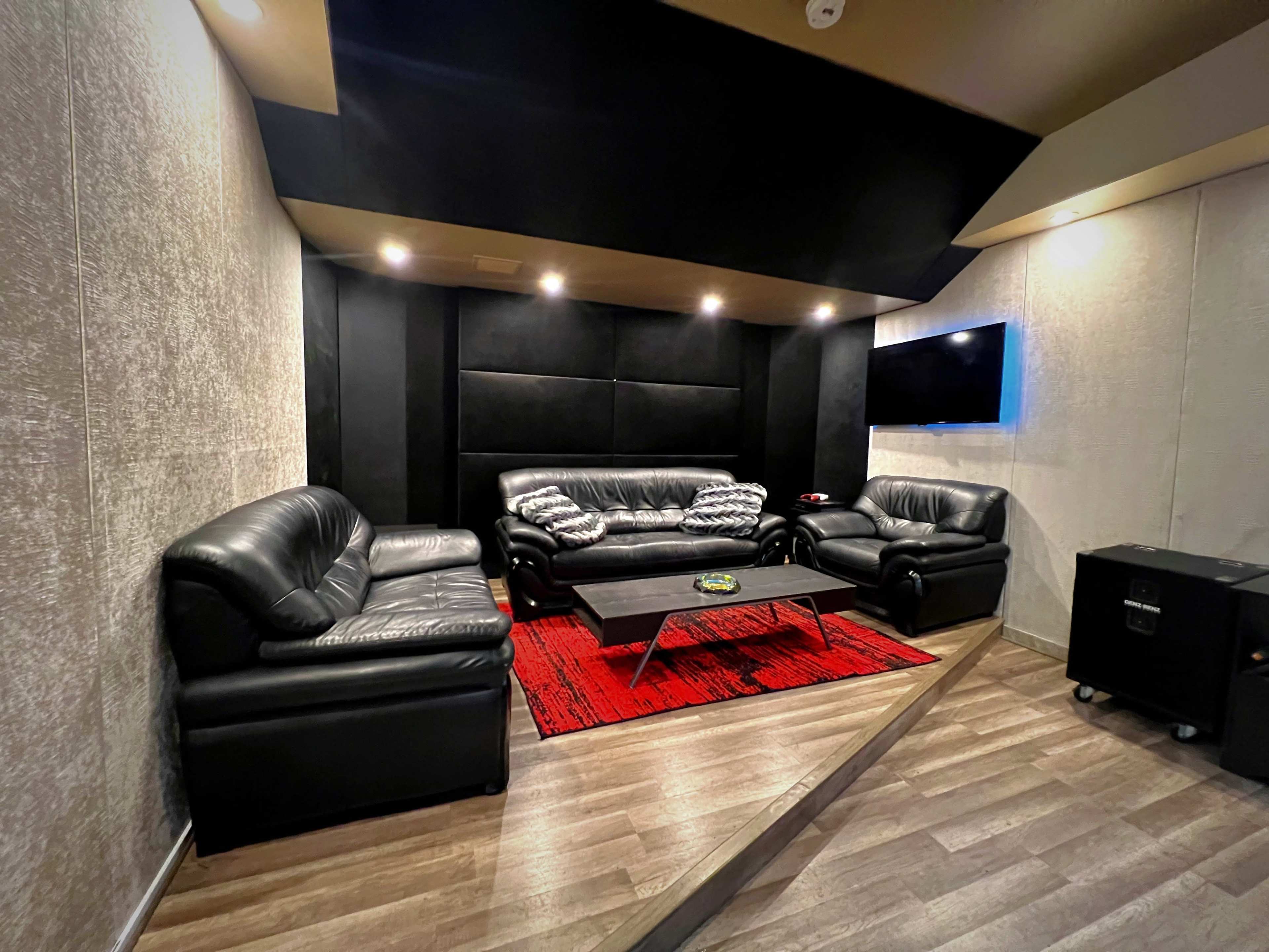 A modern lounge area with black leather sofas arranged around a coffee table on a red rug, illuminated by recessed lighting and featuring a wall-mounted television.