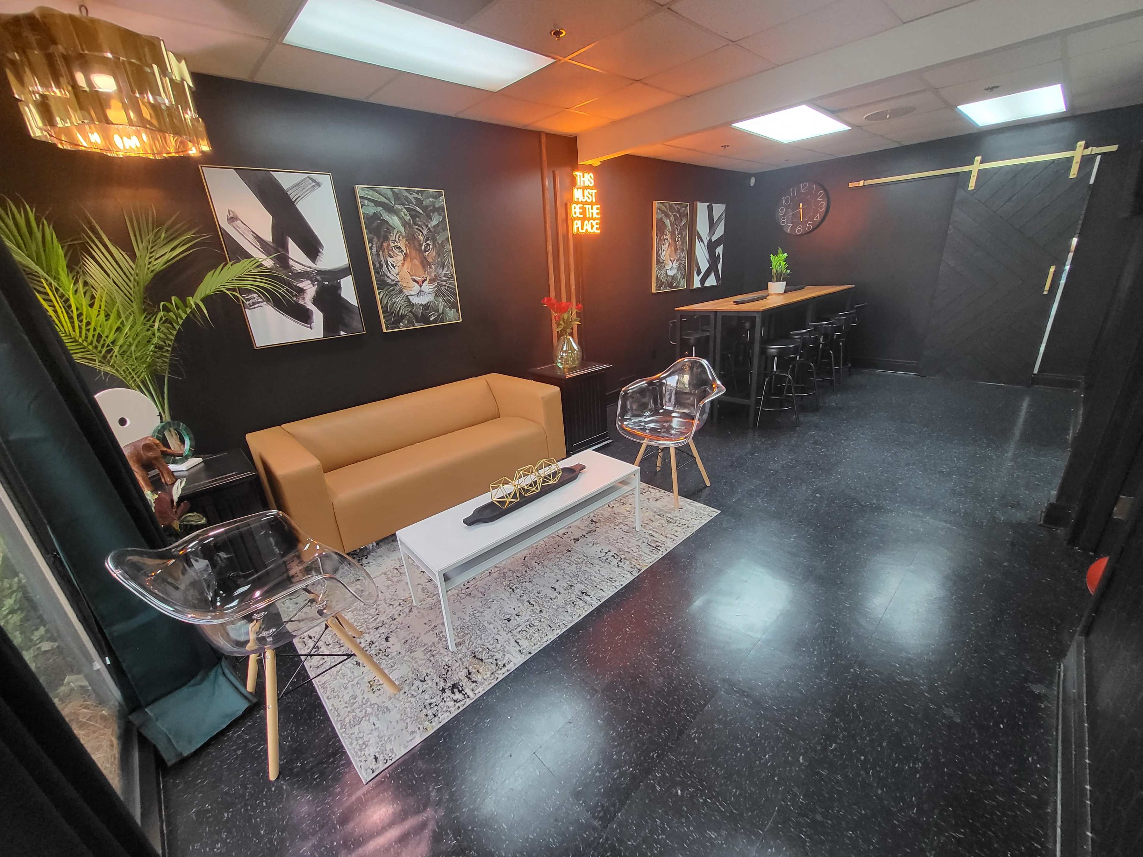 A modern lounge area with a brown leather sofa, transparent chairs, a white coffee table, and a black bar with tall stools against a dark wall, decorated with abstract artwork and greenery.