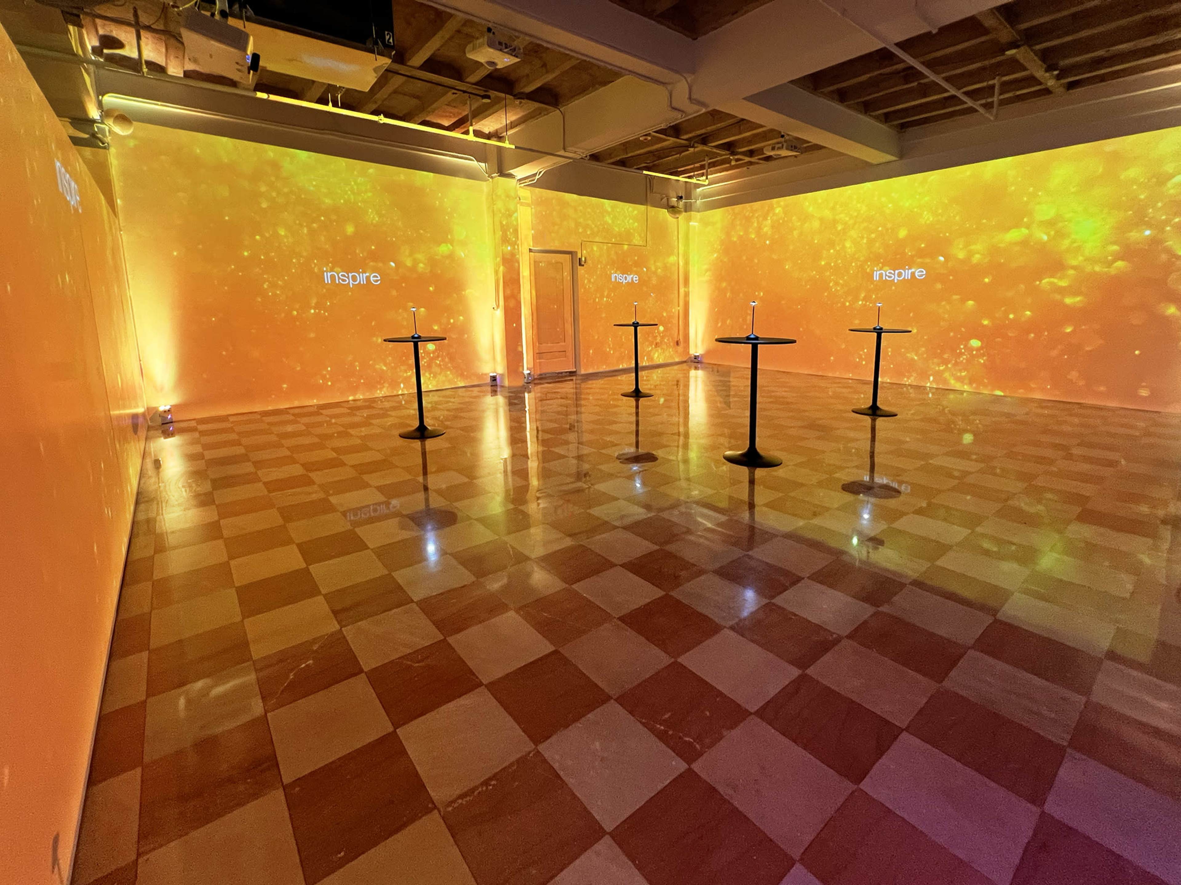 A spacious room features orange-hued walls with projection displays and several black cocktail tables arranged on a checkered floor.
