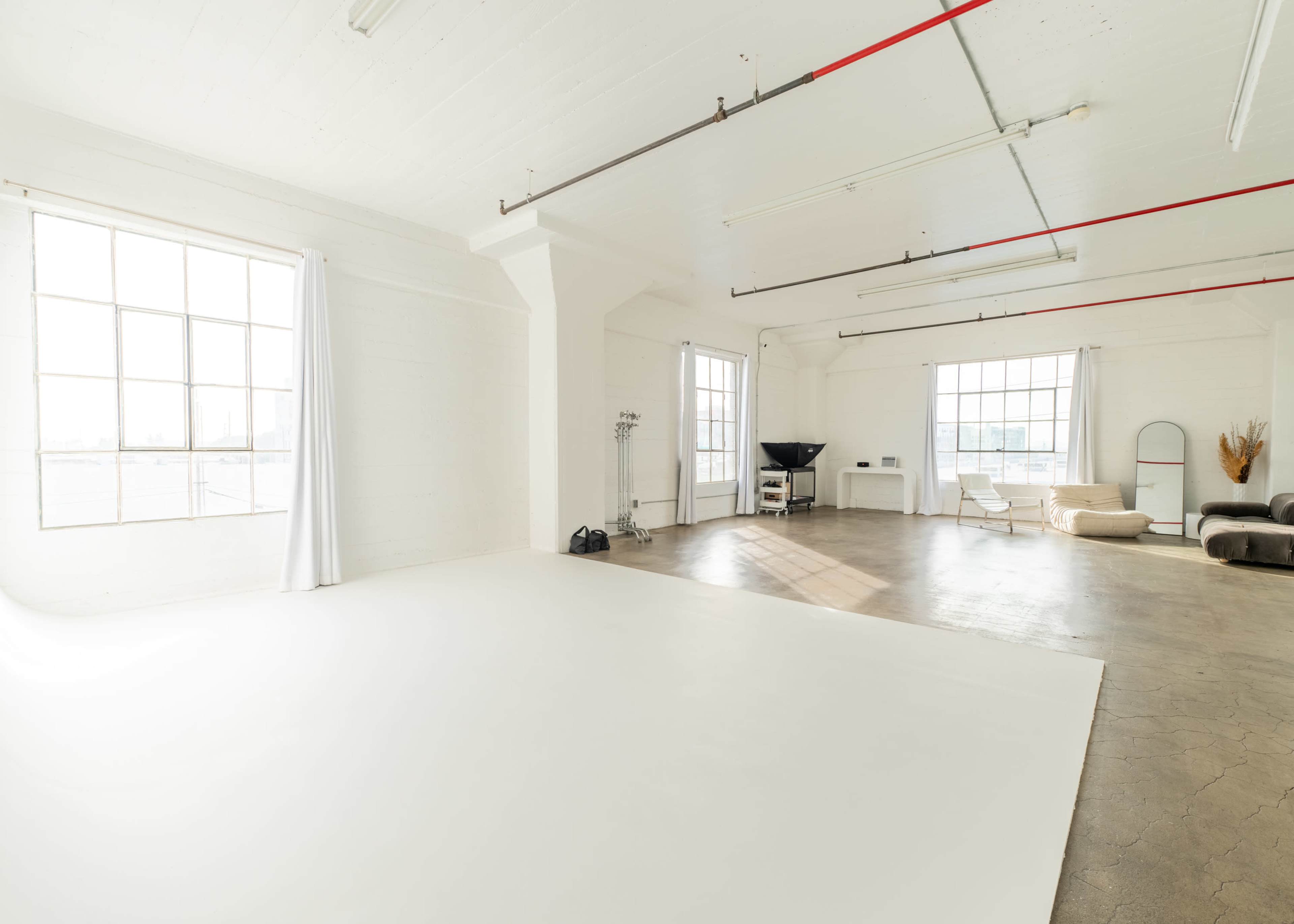 The image shows a spacious and well-lit studio with large windows, white walls, and minimalist furnishings.