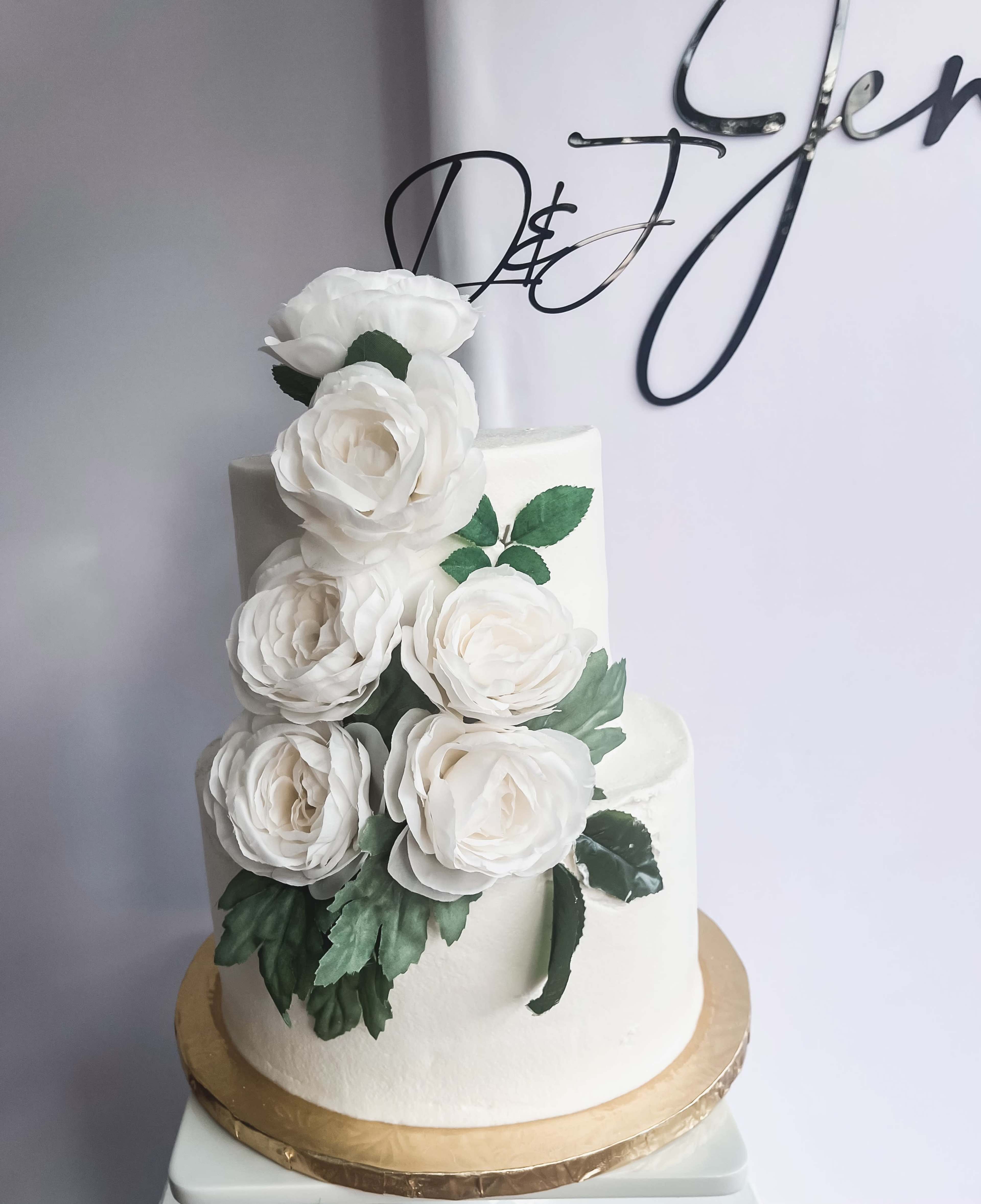 A two-tier cake adorned with white sugar roses and green leaves sits on a gold cake board against a white background.
