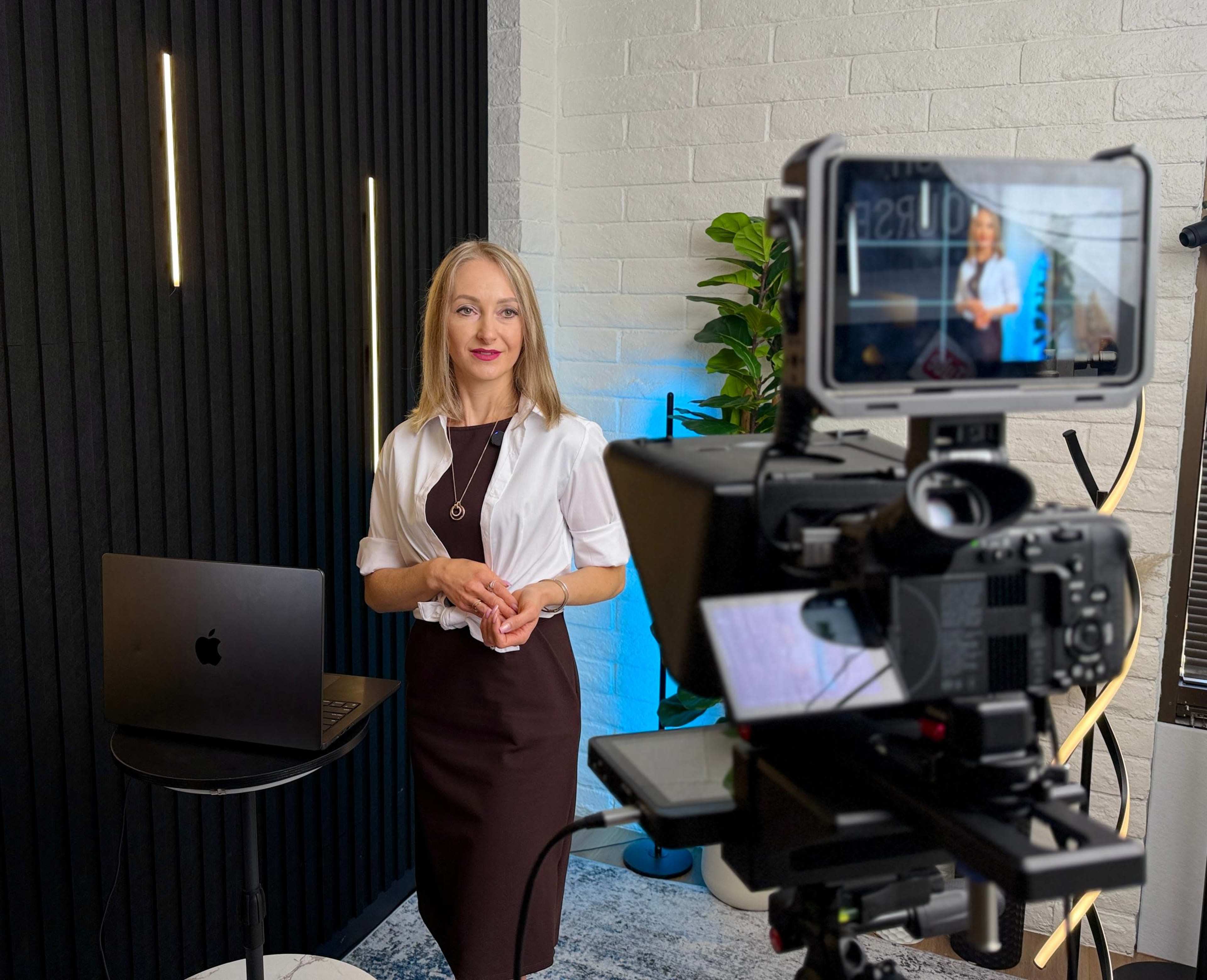 A woman stands beside a laptop and a camera setup in a modern indoor setting with decorative plants and lighting.