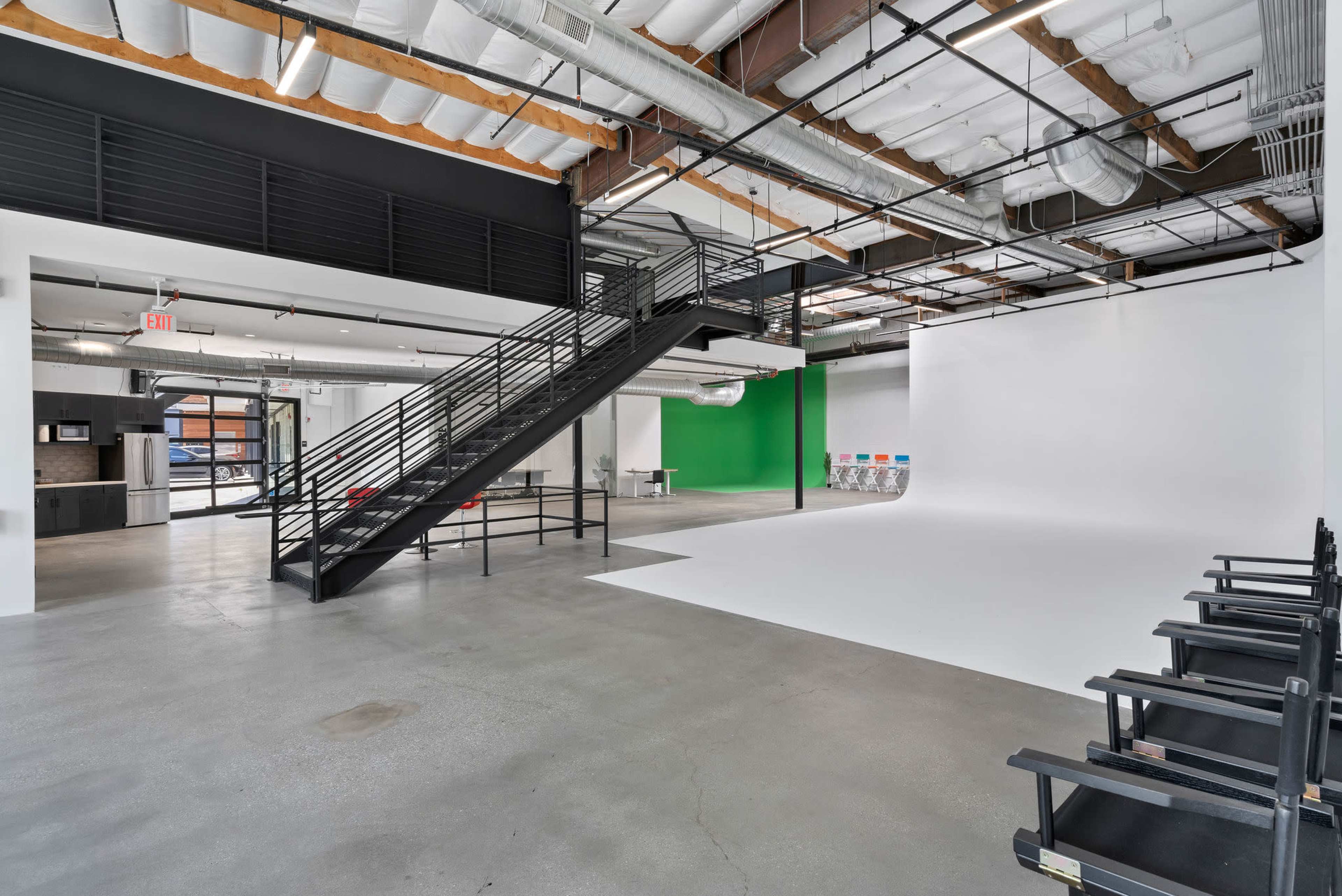 A spacious studio with a large white backdrop, a green wall, a black staircase, and several chairs arranged in front of the backdrop.
