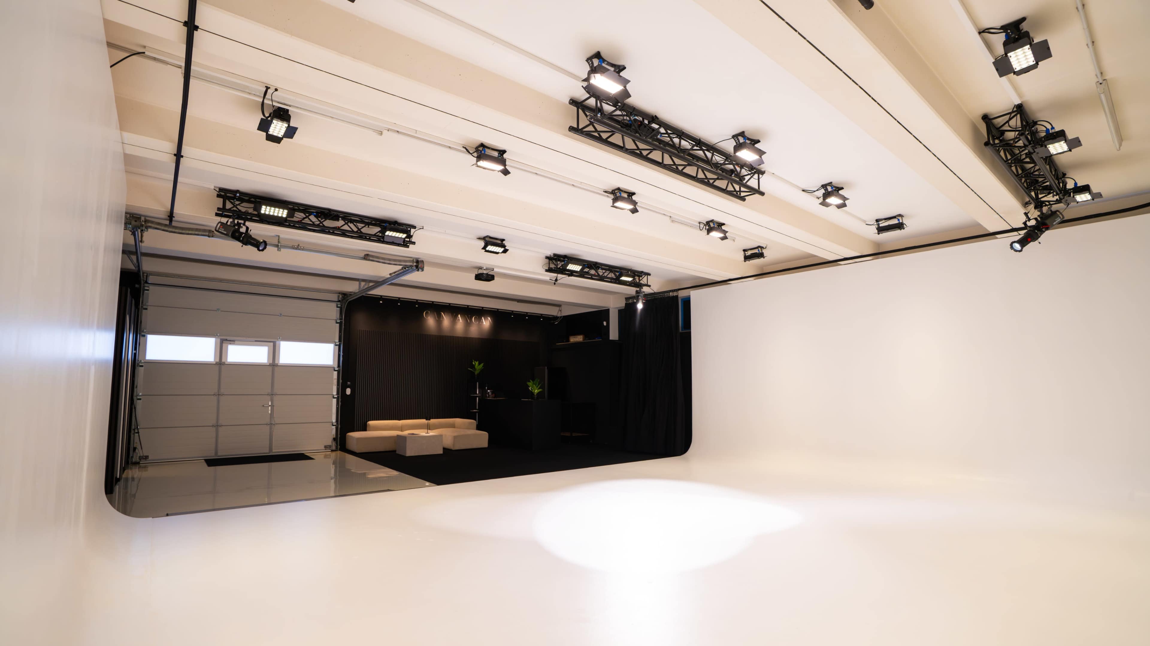 The image shows a spacious, well-lit photography studio featuring a seamless white backdrop and black curtains, with seating and a small area separated by a wall.