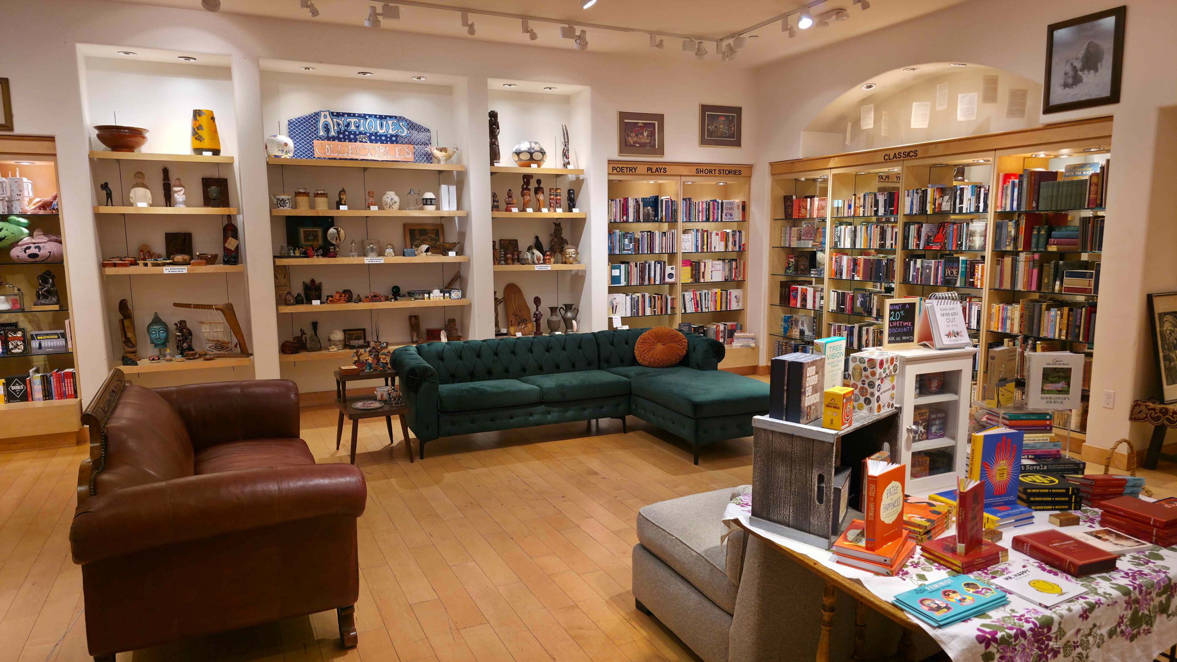 The image shows a cozy bookstore interior with bookshelves filled with various genres, a seating area featuring a green couch and brown leather chairs, and decorative items displayed on shelves.