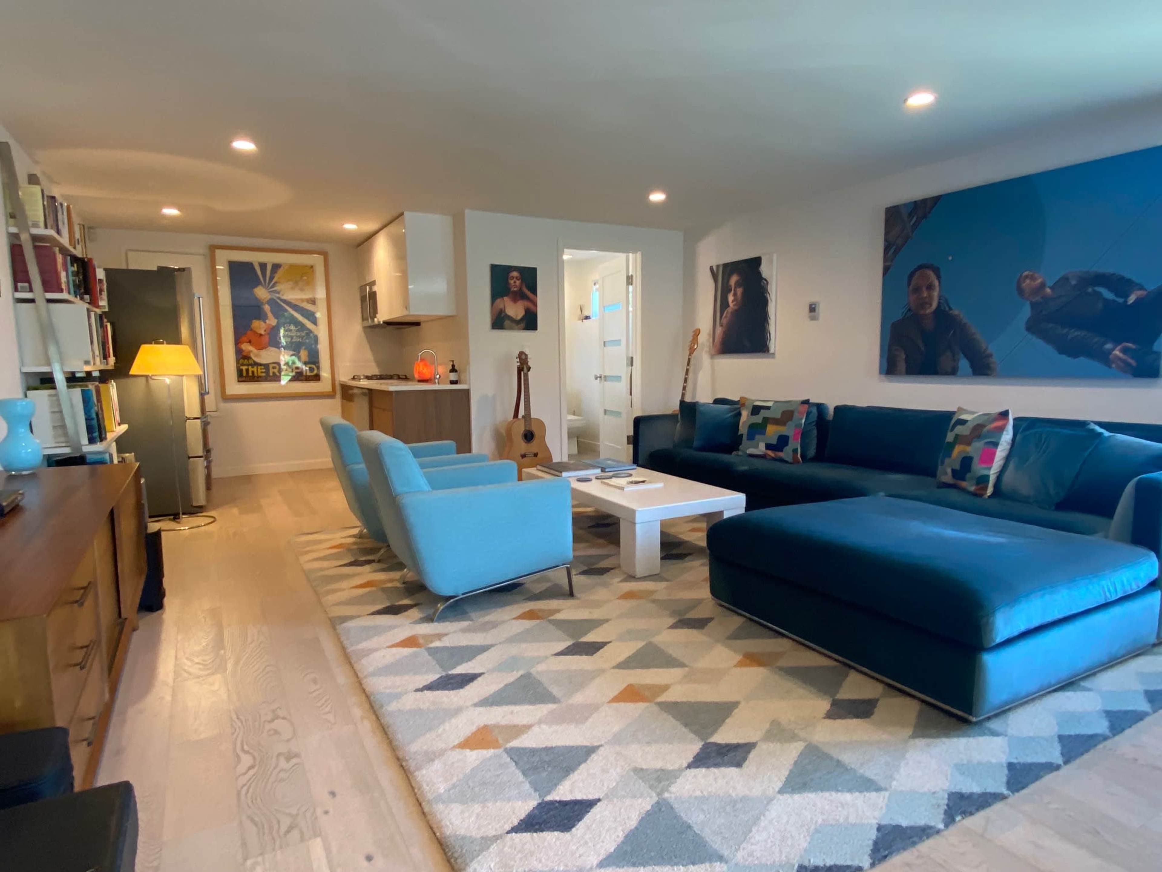 The living room features a blue sectional sofa, a coffee table, a patterned rug, and various artworks on the walls, with a bookshelf and a guitar nearby.