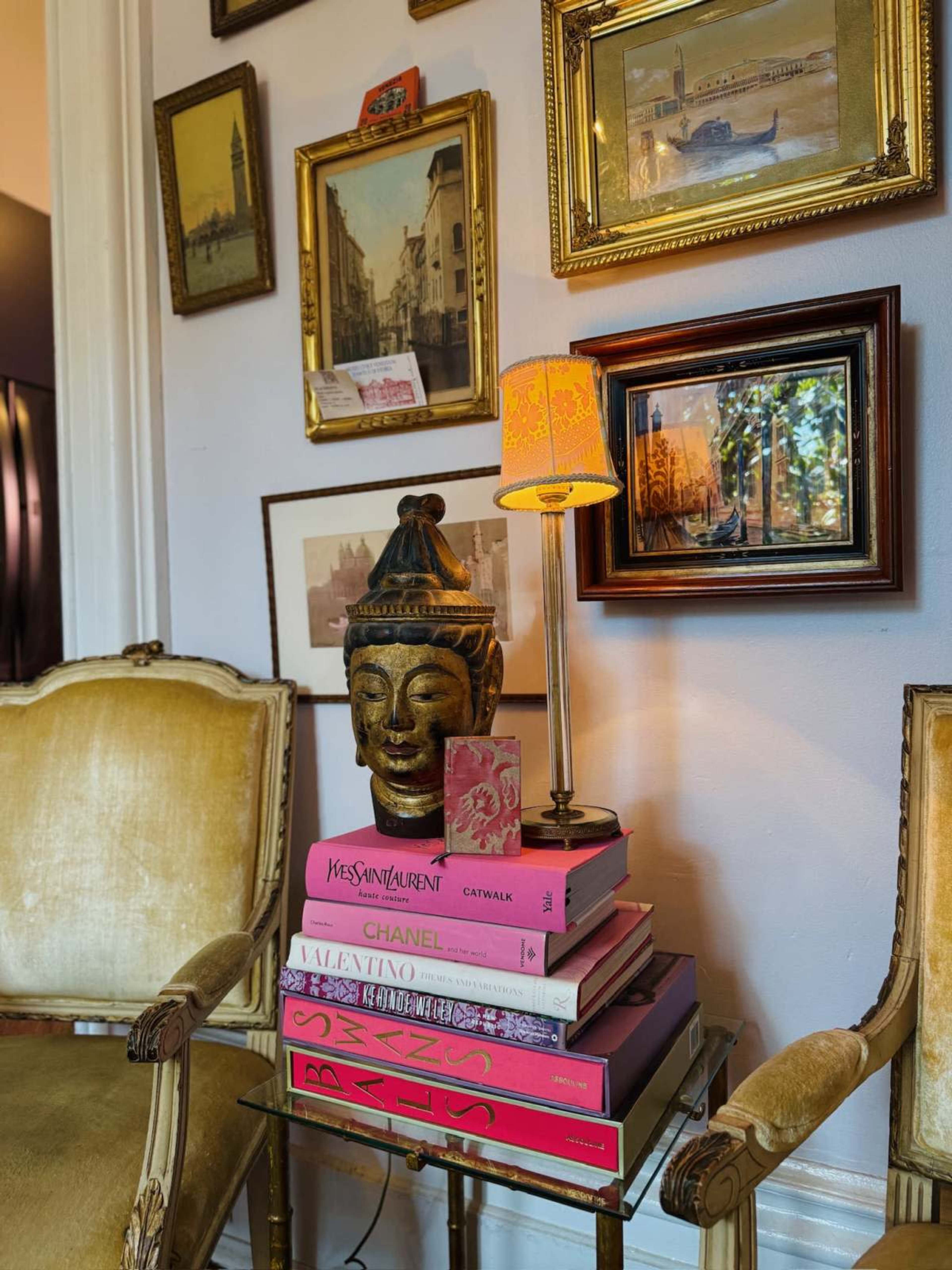 A sculpted Buddha head sits atop a stack of colorful fashion books on a glass table, illuminated by a small lamp, while framed art hangs on the wall behind.
