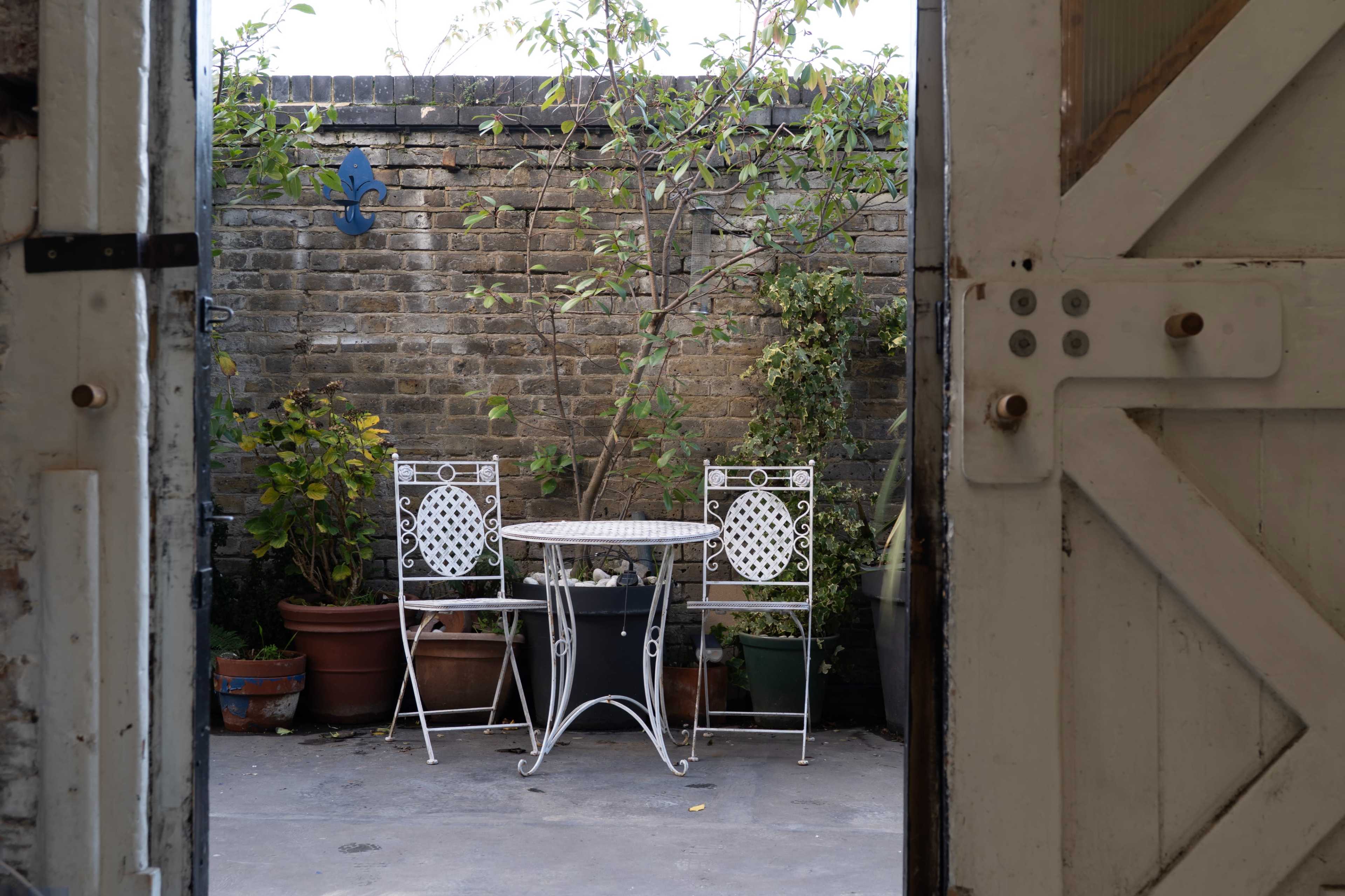 A small outdoor patio features a round table surrounded by two white metal chairs, with potted plants and a brick wall in the background.