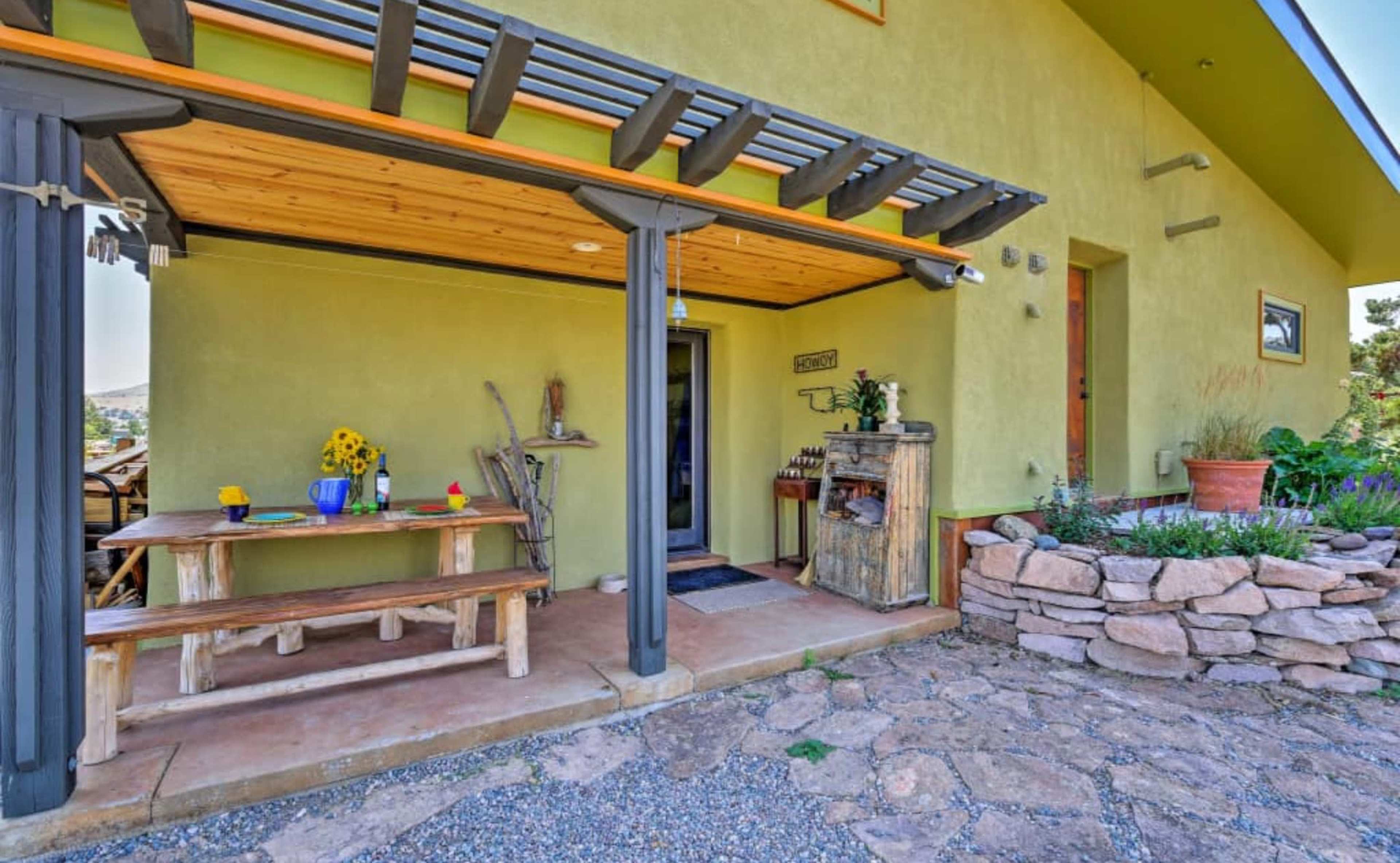 A small porch area with a wooden table and chairs, a rustic side table, and potted plants beside a green exterior wall of a house.