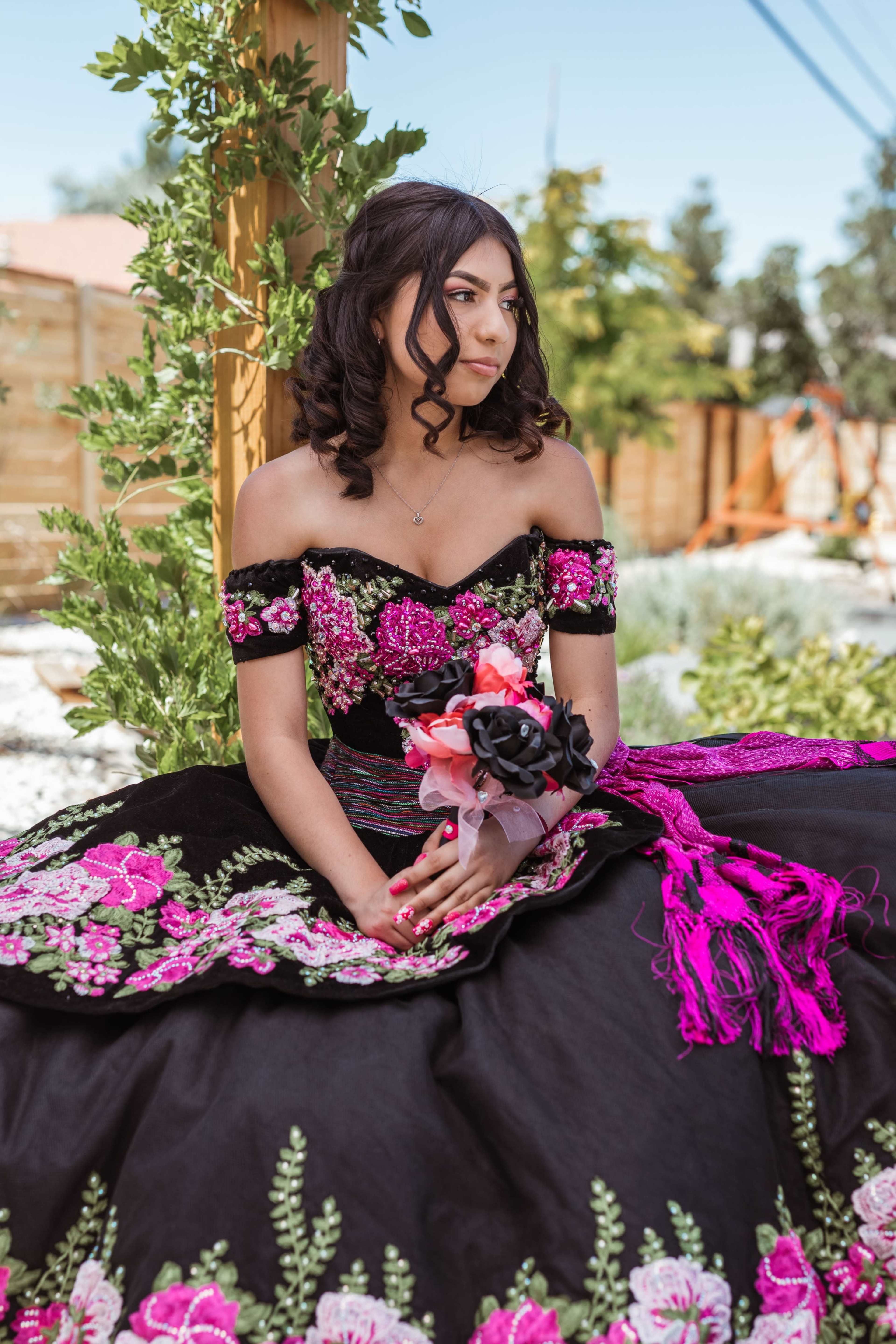 A young woman in a black traditional dress adorned with colorful floral embroidery sits holding a bouquet of roses.