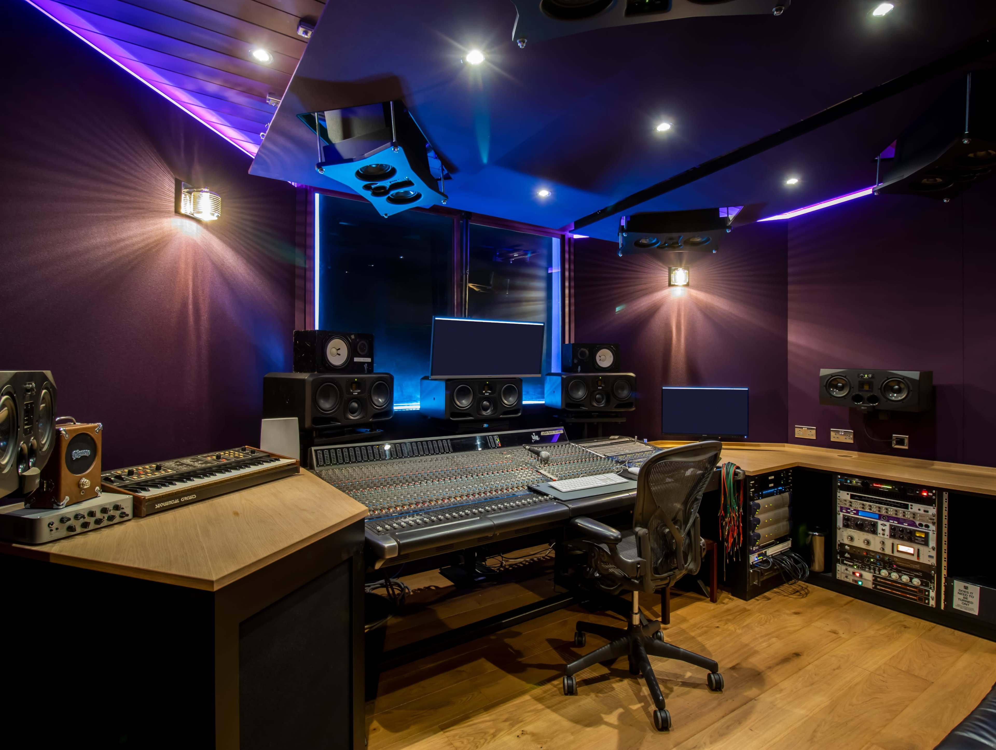 The image shows a modern music recording studio with a large mixing console, multiple speakers, and ambient lighting.