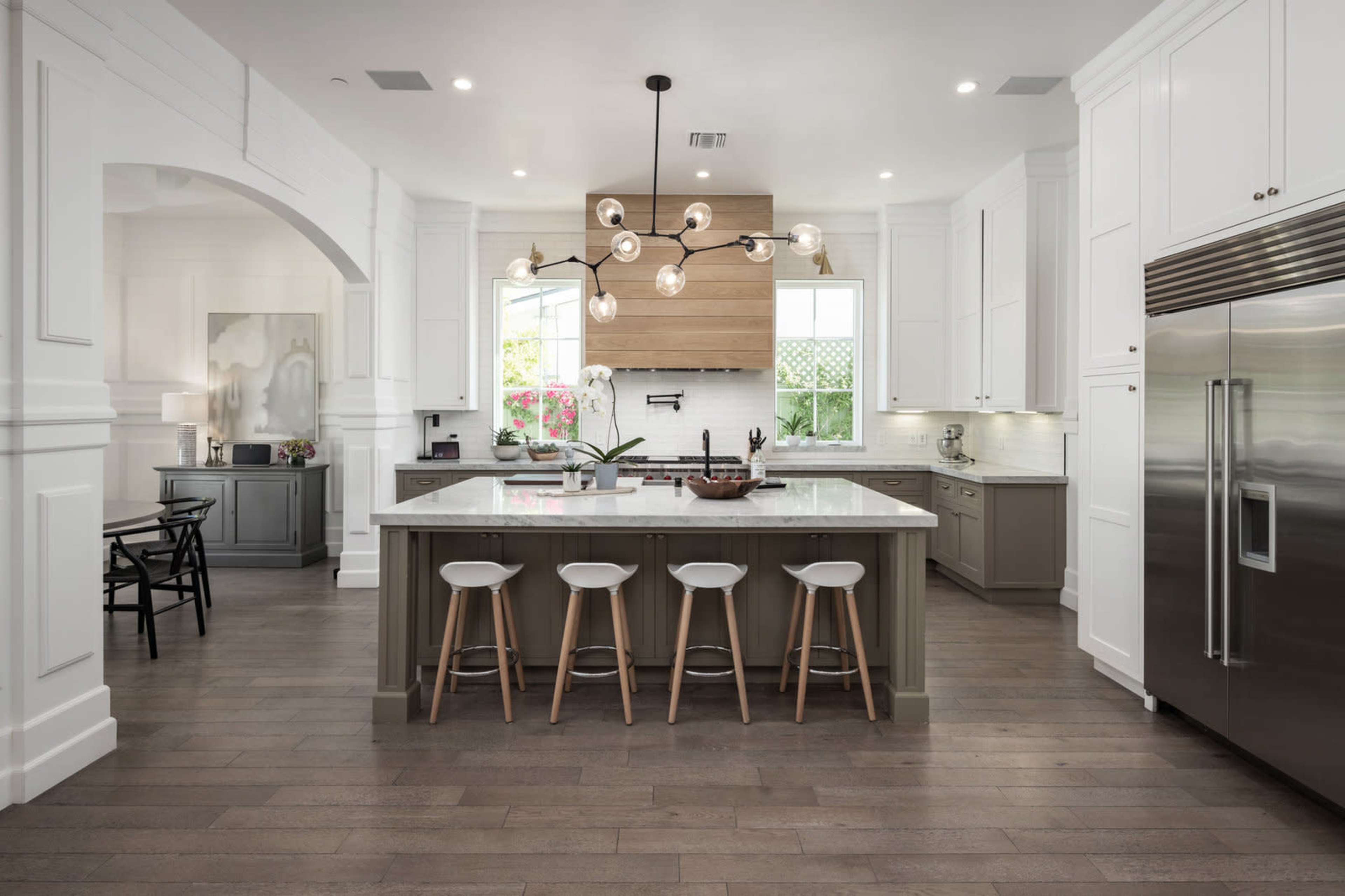 The image shows a modern kitchen with a central island, pendant lighting, and white cabinetry, featuring a mix of dark and light wood elements.