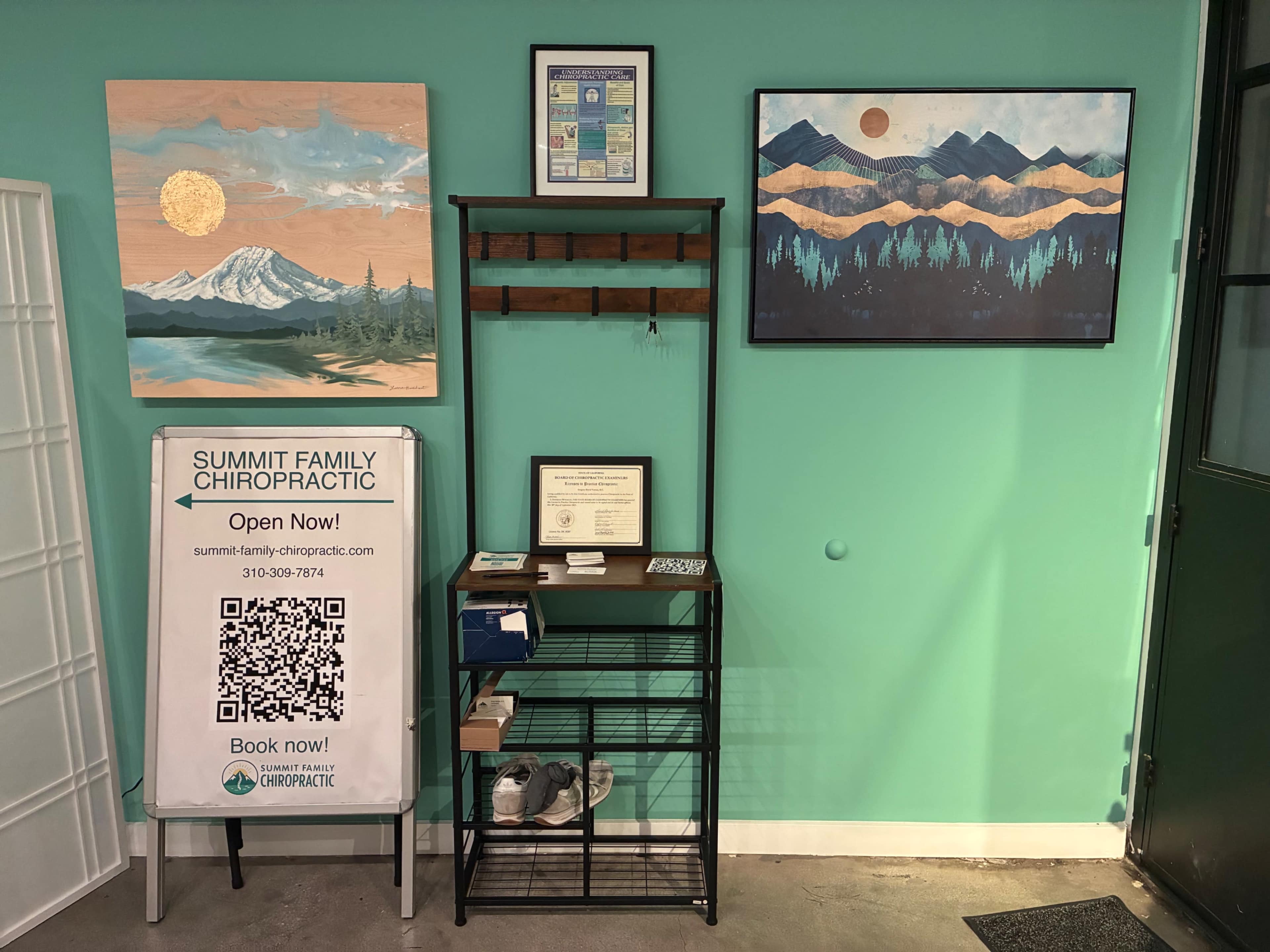 The image shows a brightly painted turquoise wall featuring two landscape paintings, alongside a black metal shelf with a sign for Summit Family Chiropractic, a QR code, and various documents displayed on the wall.