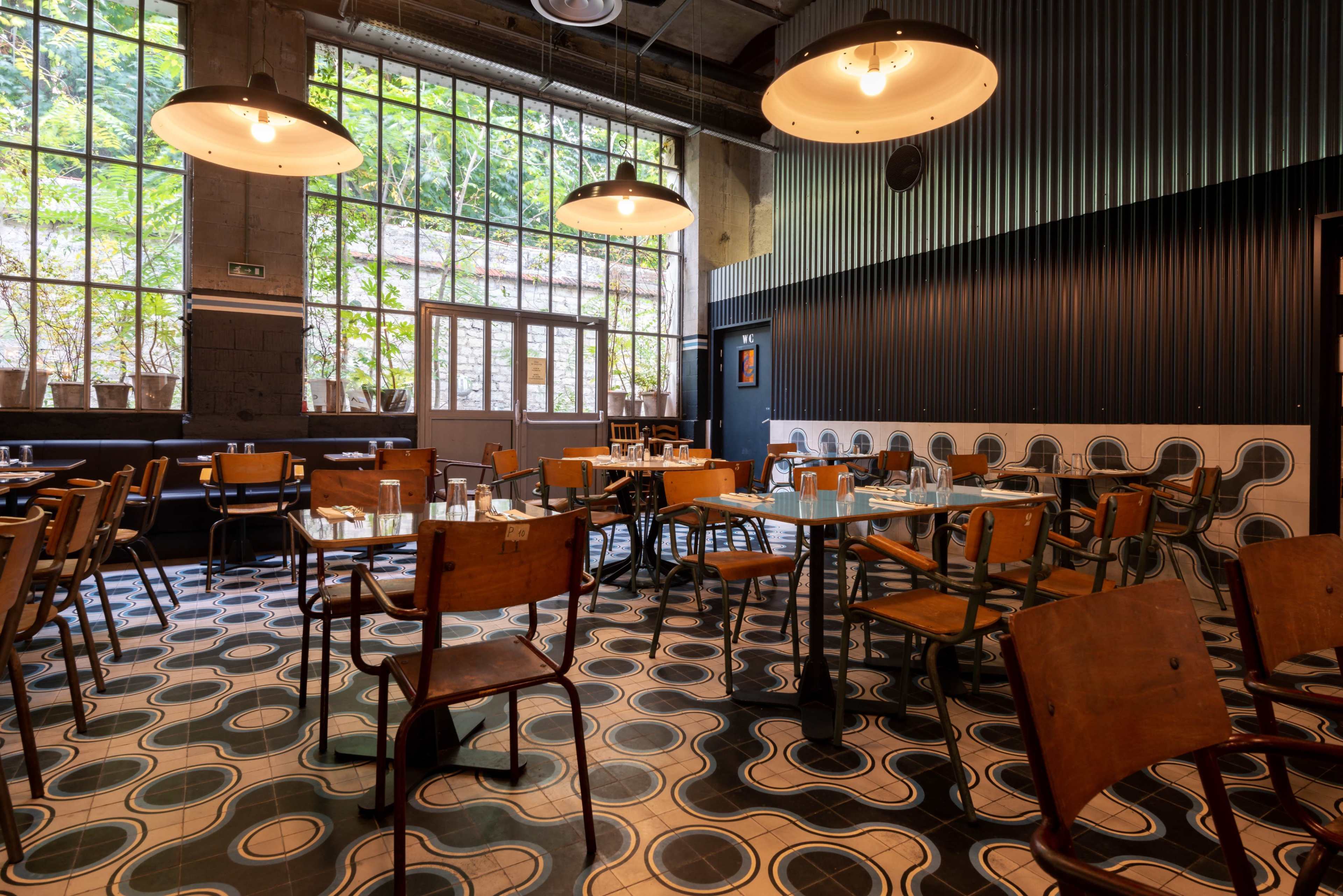 An interior of a restaurant with wooden chairs, tables, and large windows allowing natural light to enter, contrasting with the black and white patterned tile floor.