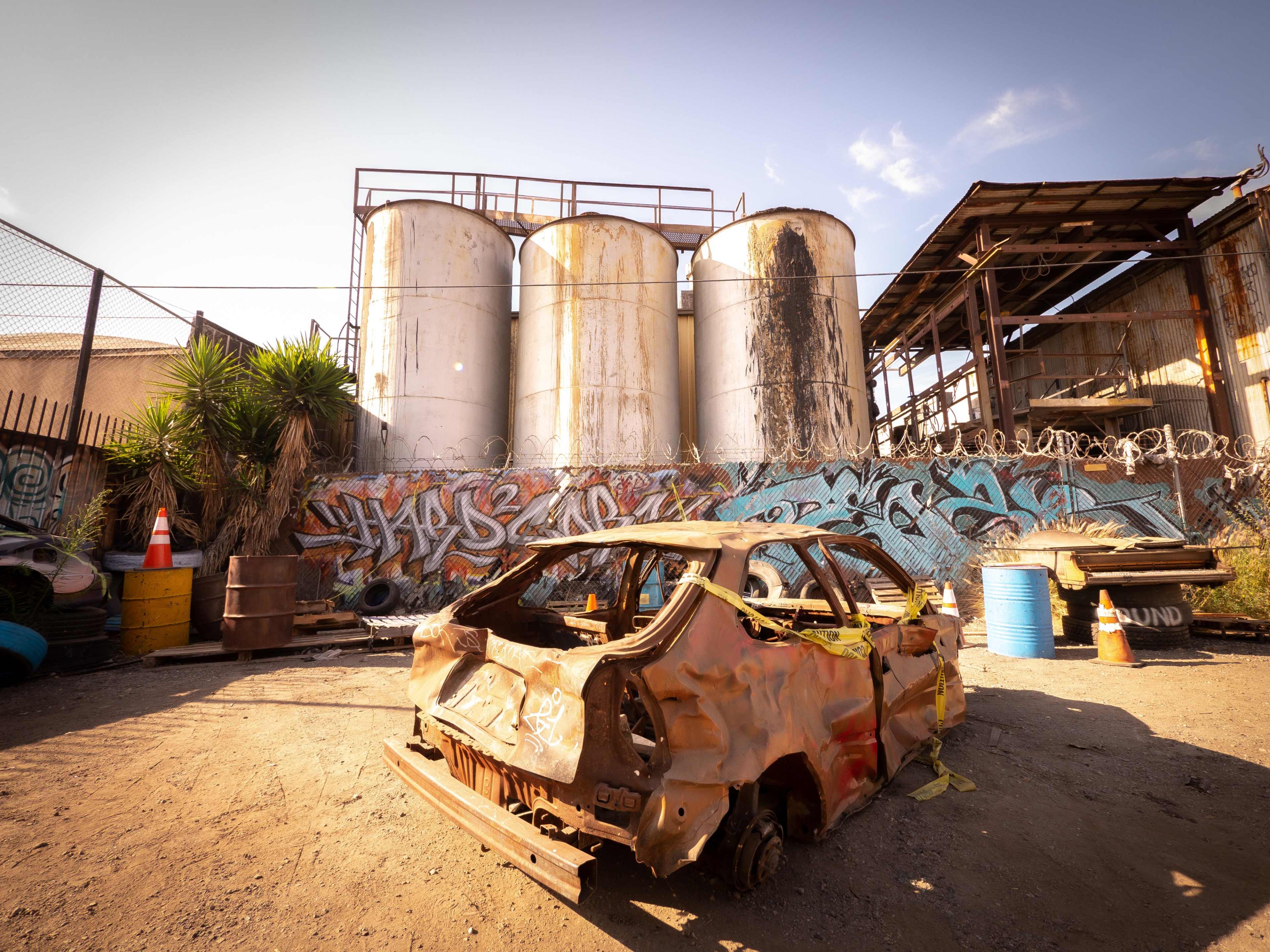 The image shows a rusted, abandoned car in front of large, weathered storage tanks surrounded by graffiti-covered walls.