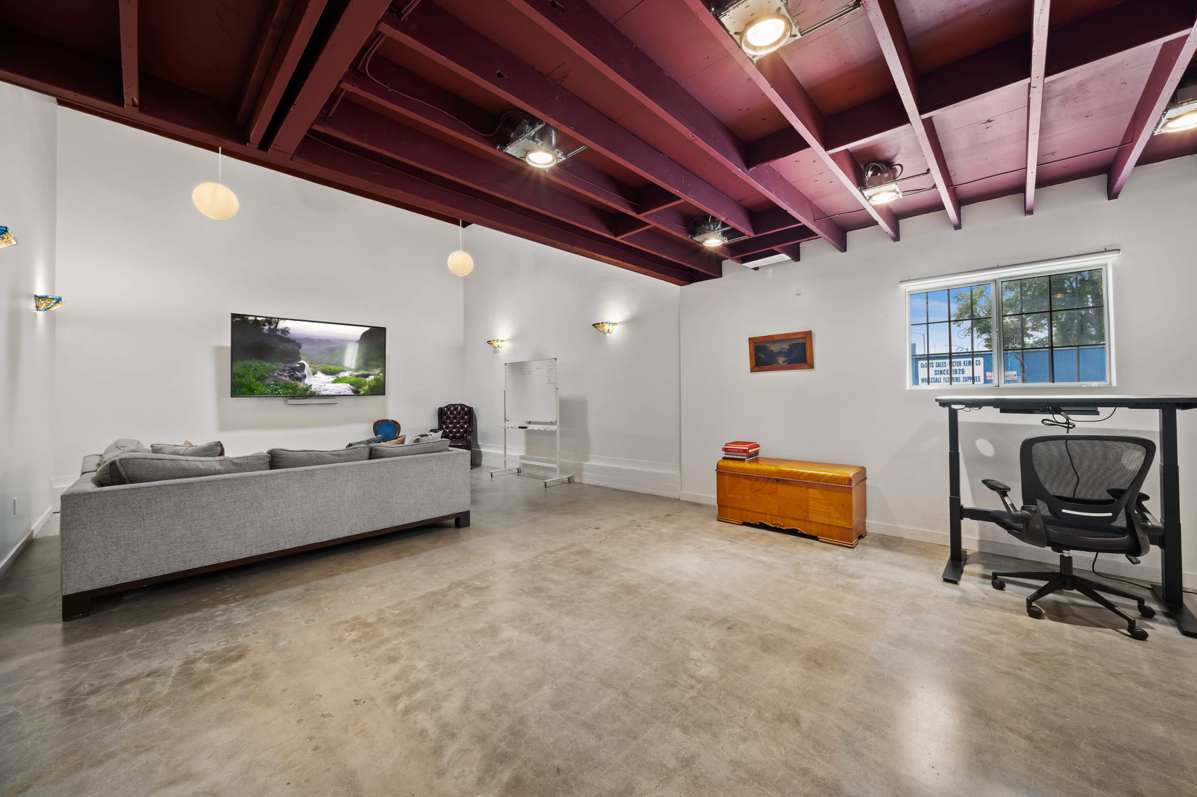 The image shows a modern interior space featuring a gray sofa, a wooden chest, a desk with an ergonomic chair, and a wall-mounted television.