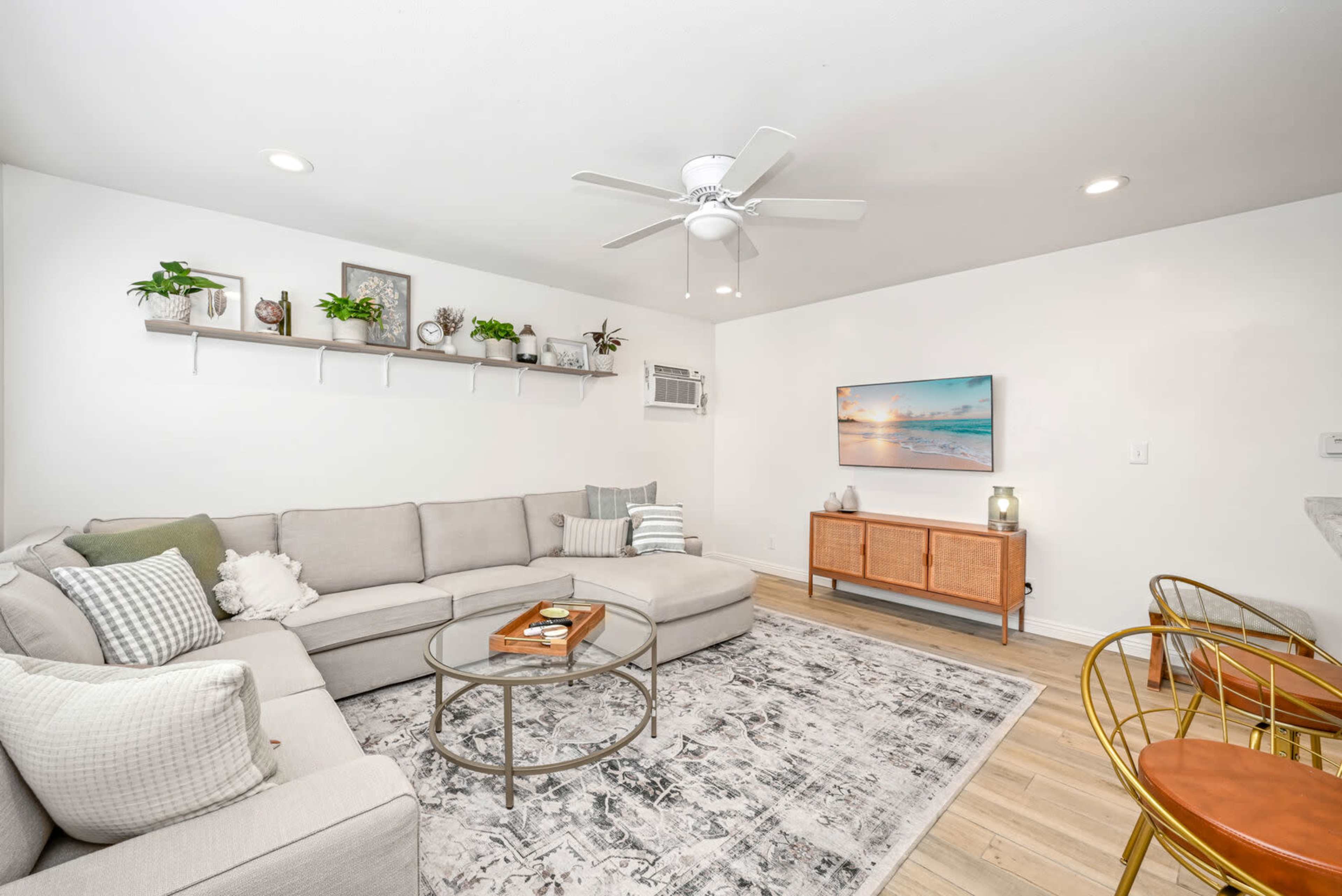A modern living room features a sectional sofa, coffee table, and a TV mounted on the wall, with plants on a shelf and wooden furniture.