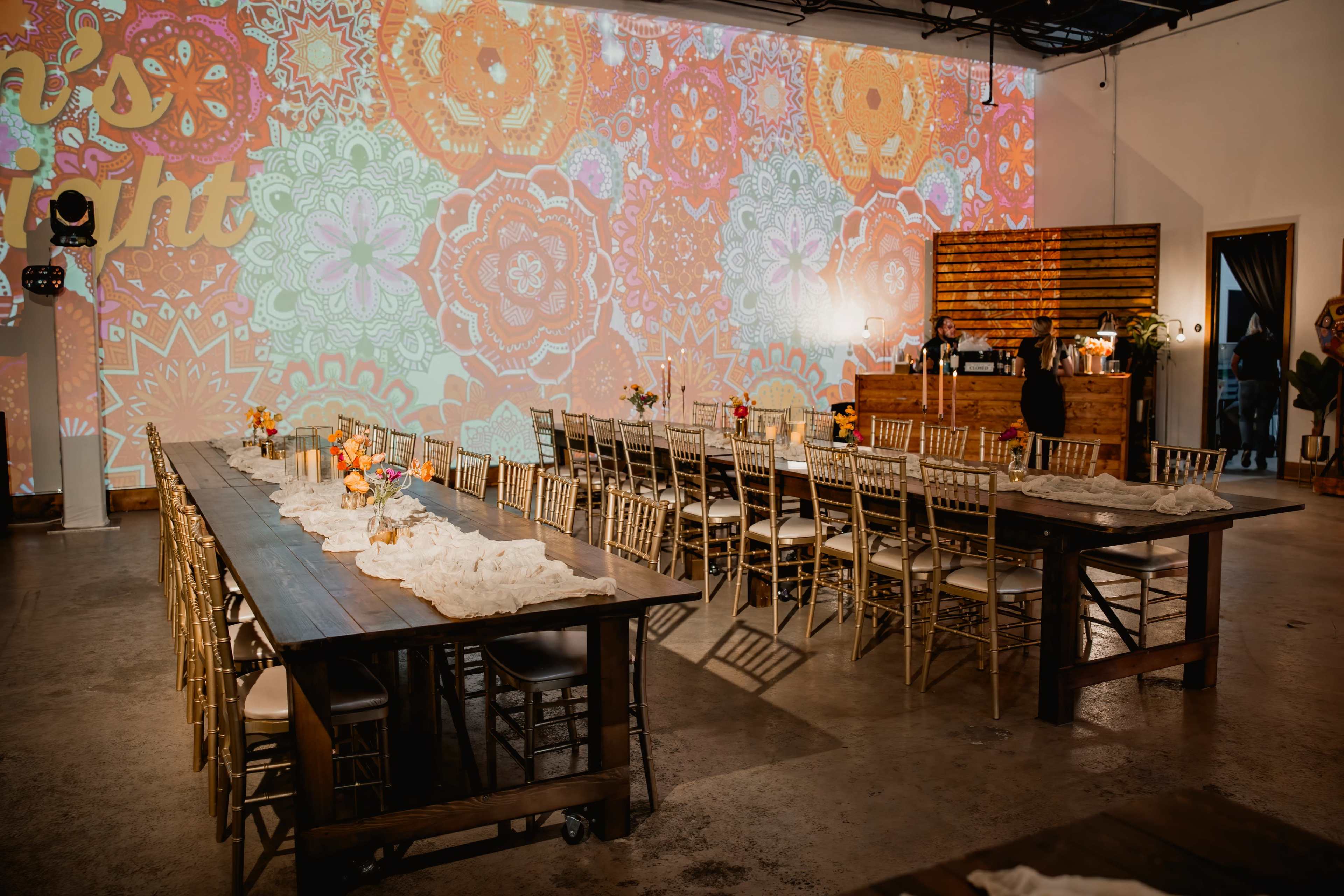A spacious event venue features long wooden tables set for a gathering, with vibrant patterned projections illuminating the walls.