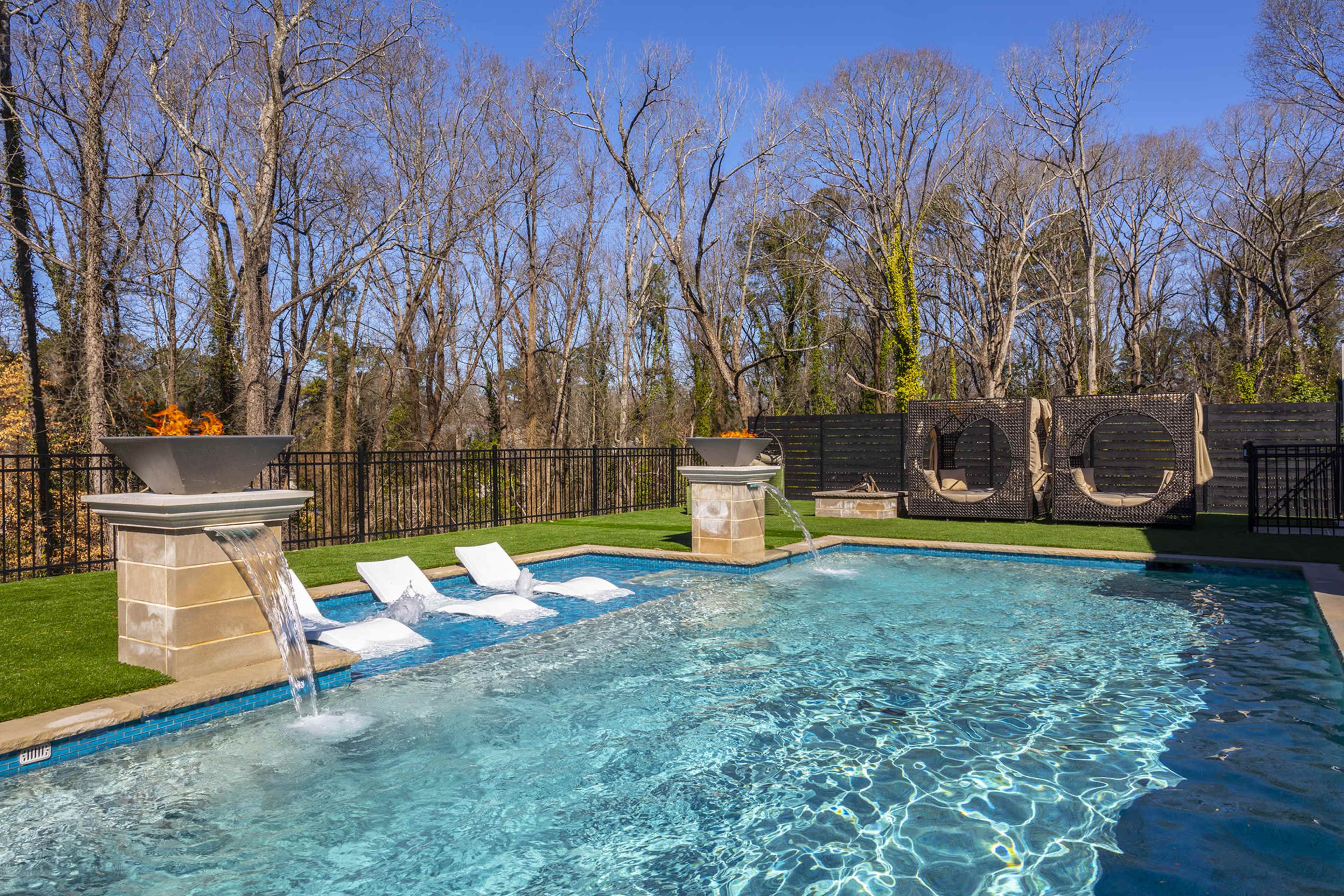 A residential outdoor pool surrounded by landscaped grass, with lounge chairs and a stone wall in the background.