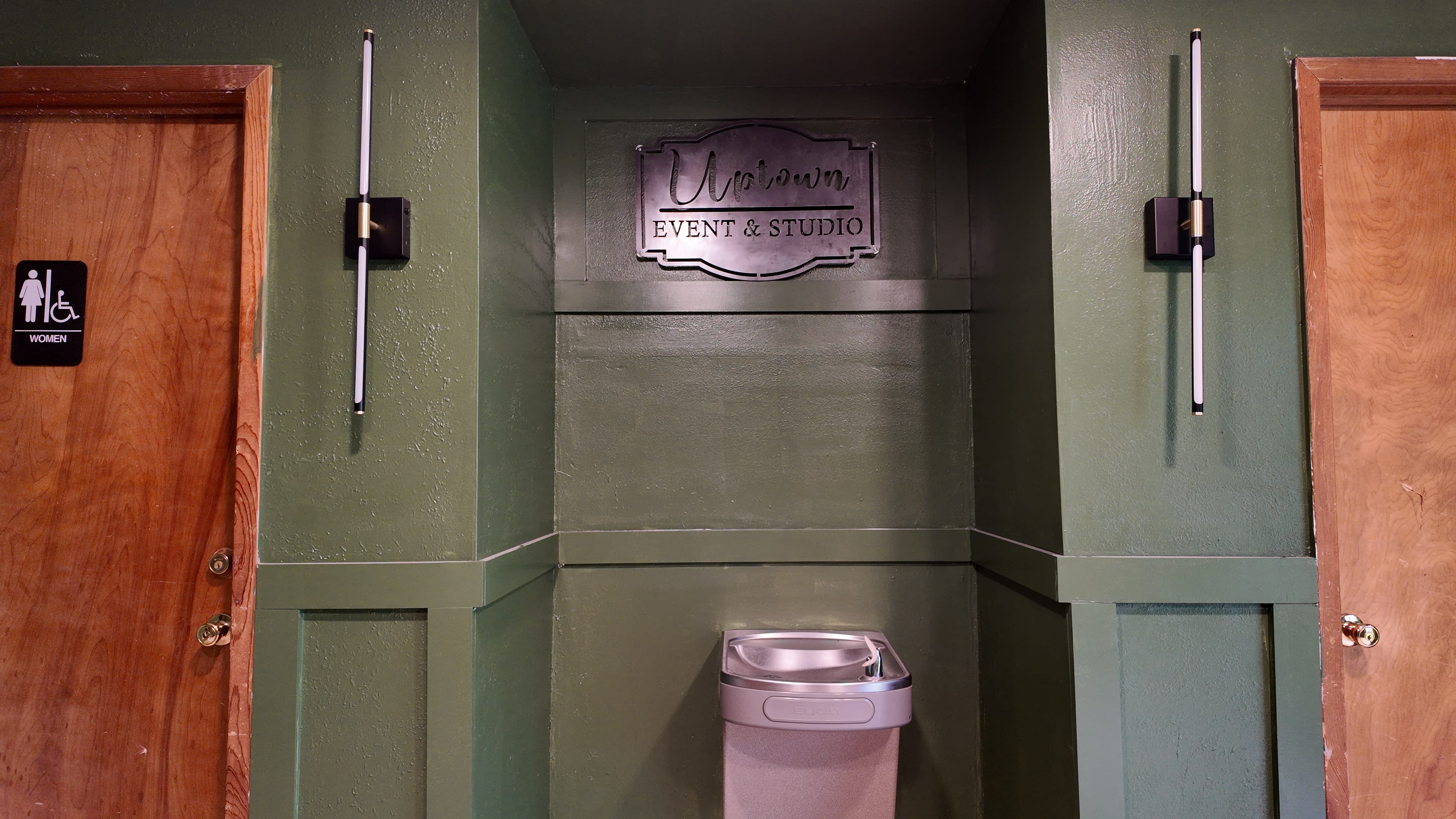 The image shows a hallway with a sign that reads "Uritarium Event & Studio" above a water fountain and next to a wooden door labeled "WOMEN."