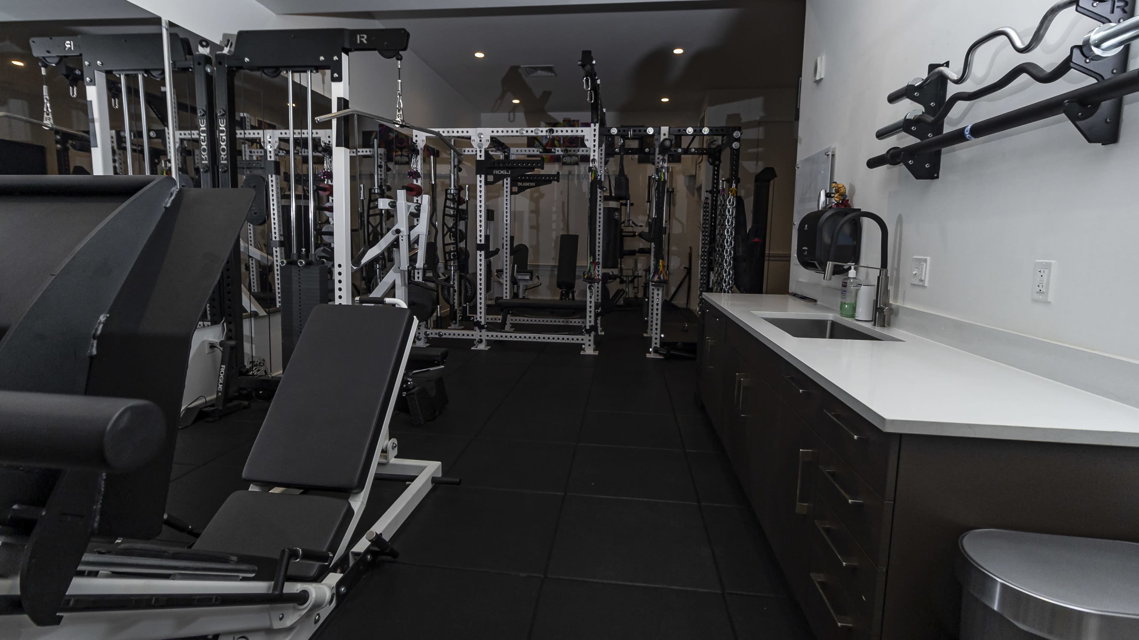 A modern gym interior with various weightlifting equipment, a bench press, and a small kitchenette area.