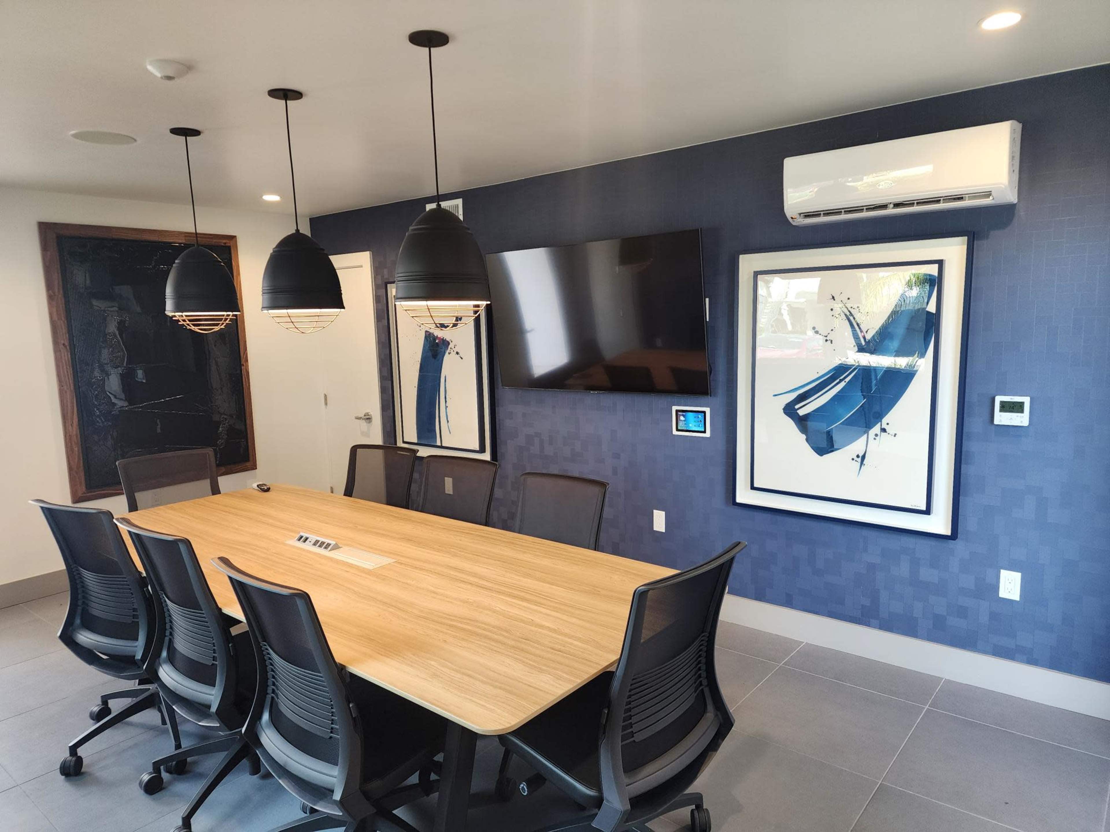 A modern conference room features a large wooden table surrounded by six black chairs, with two framed artworks and a wall-mounted television.