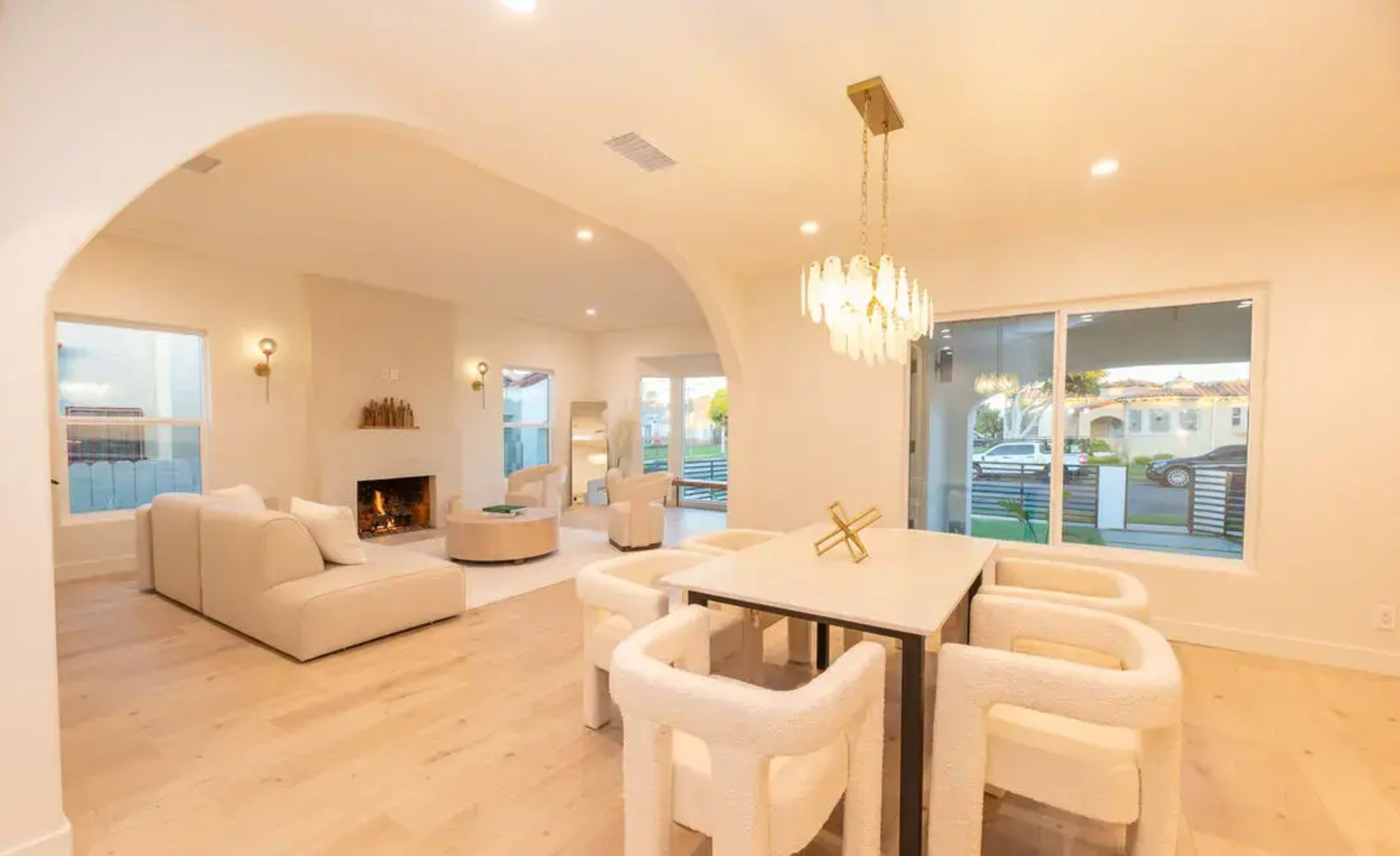 The image shows a modern living room and dining area with a fireplace, contemporary furniture, and large windows illuminating the space.