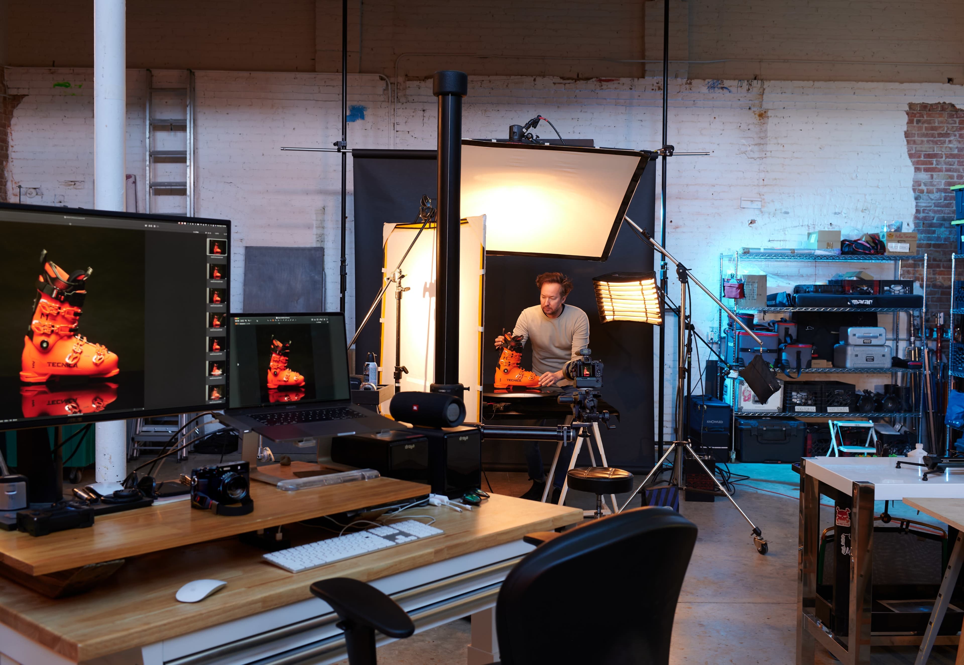 A photography studio is set up with a photographer working in front of a backdrop, surrounded by equipment and computer screens displaying images of ski boots.