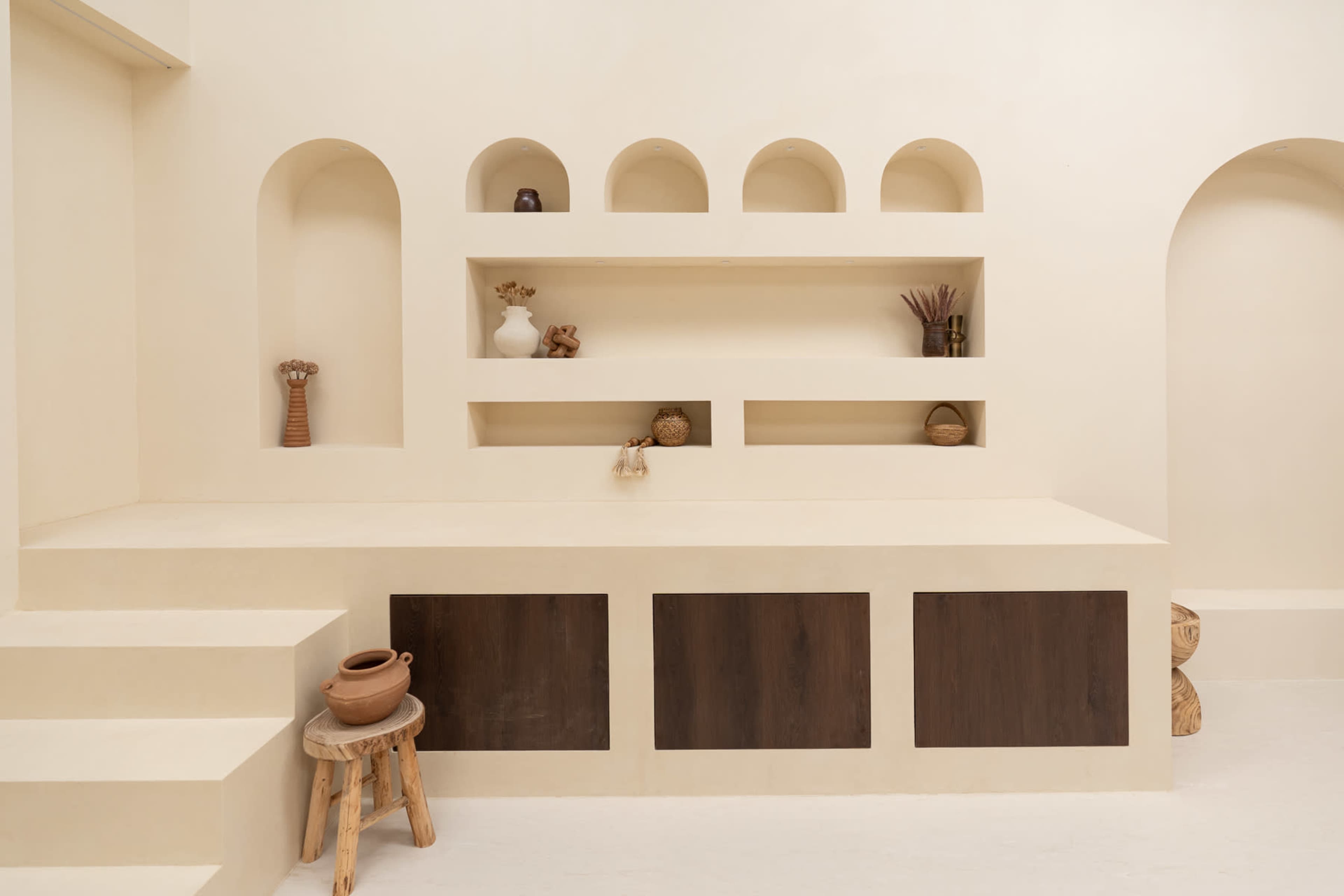 A minimalist interior features a cream-colored wall with curved shelves displaying decorative items, accompanied by a wooden cabinet and a small stool.