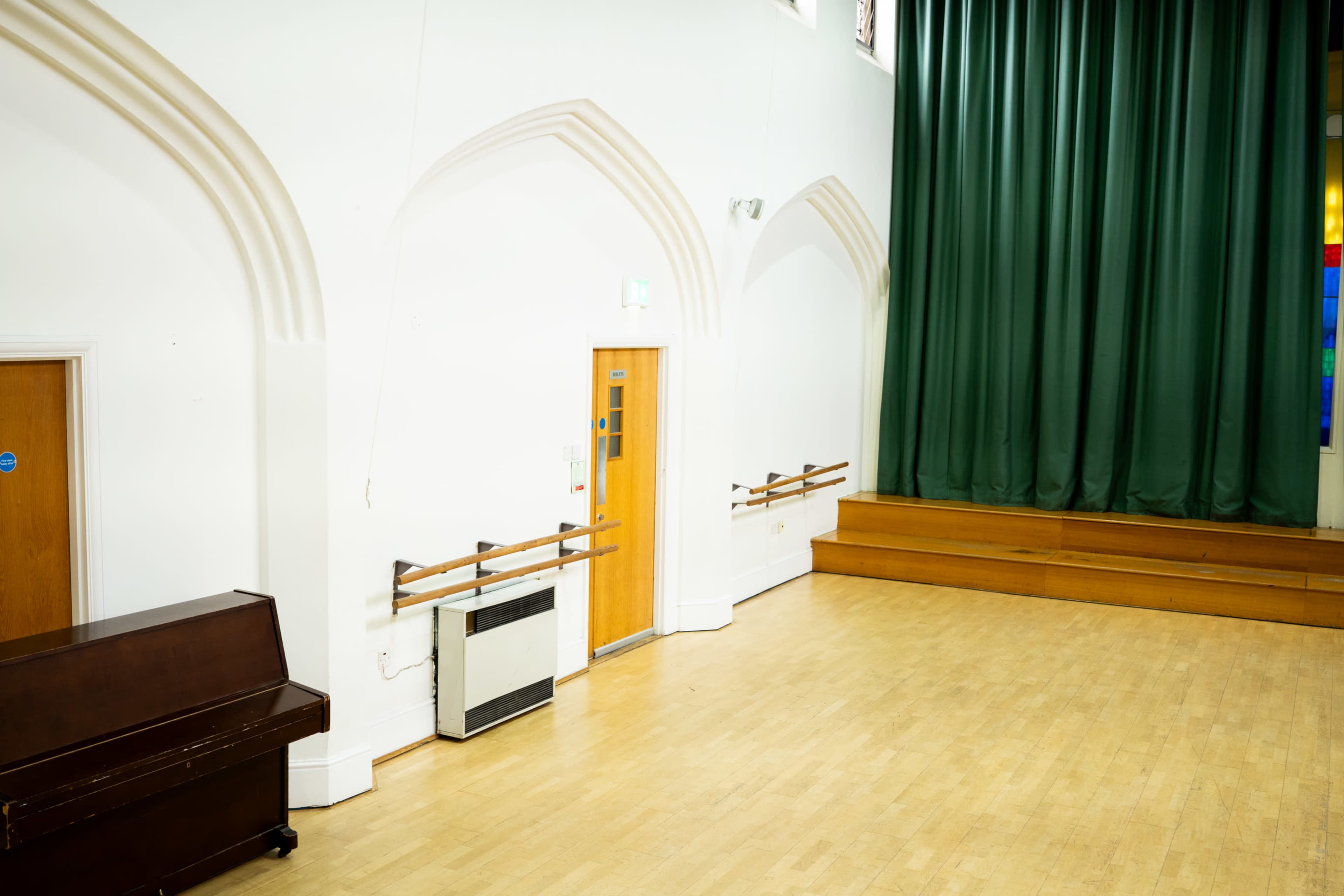 The image shows a spacious, empty room with wooden flooring, a large green curtain at one end, a piano against one wall, and two doors with wall-mounted ballet bars beside them.