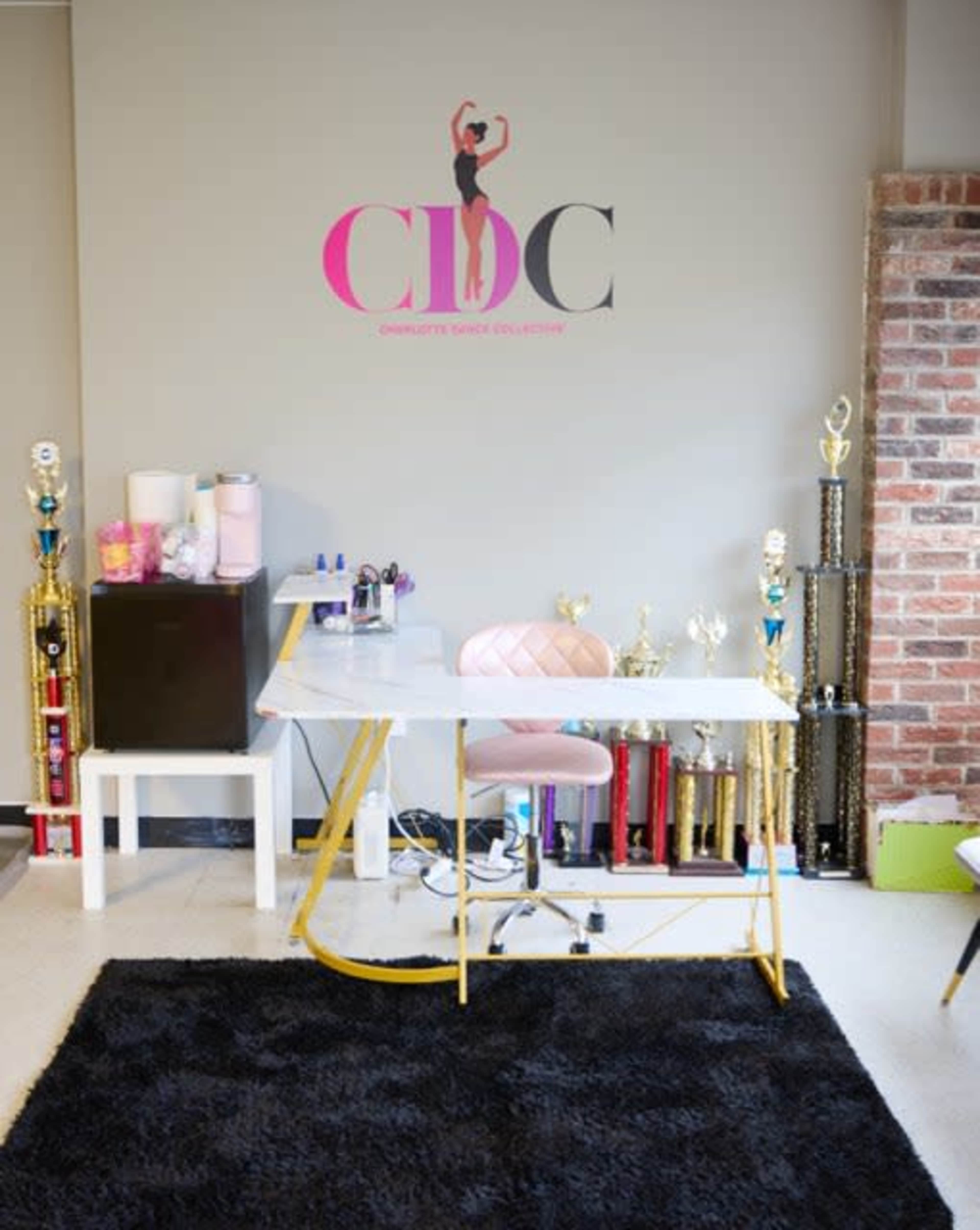 A dance studio workspace features a white desk with a pink chair, surrounded by shelves filled with trophies and a soft black rug.