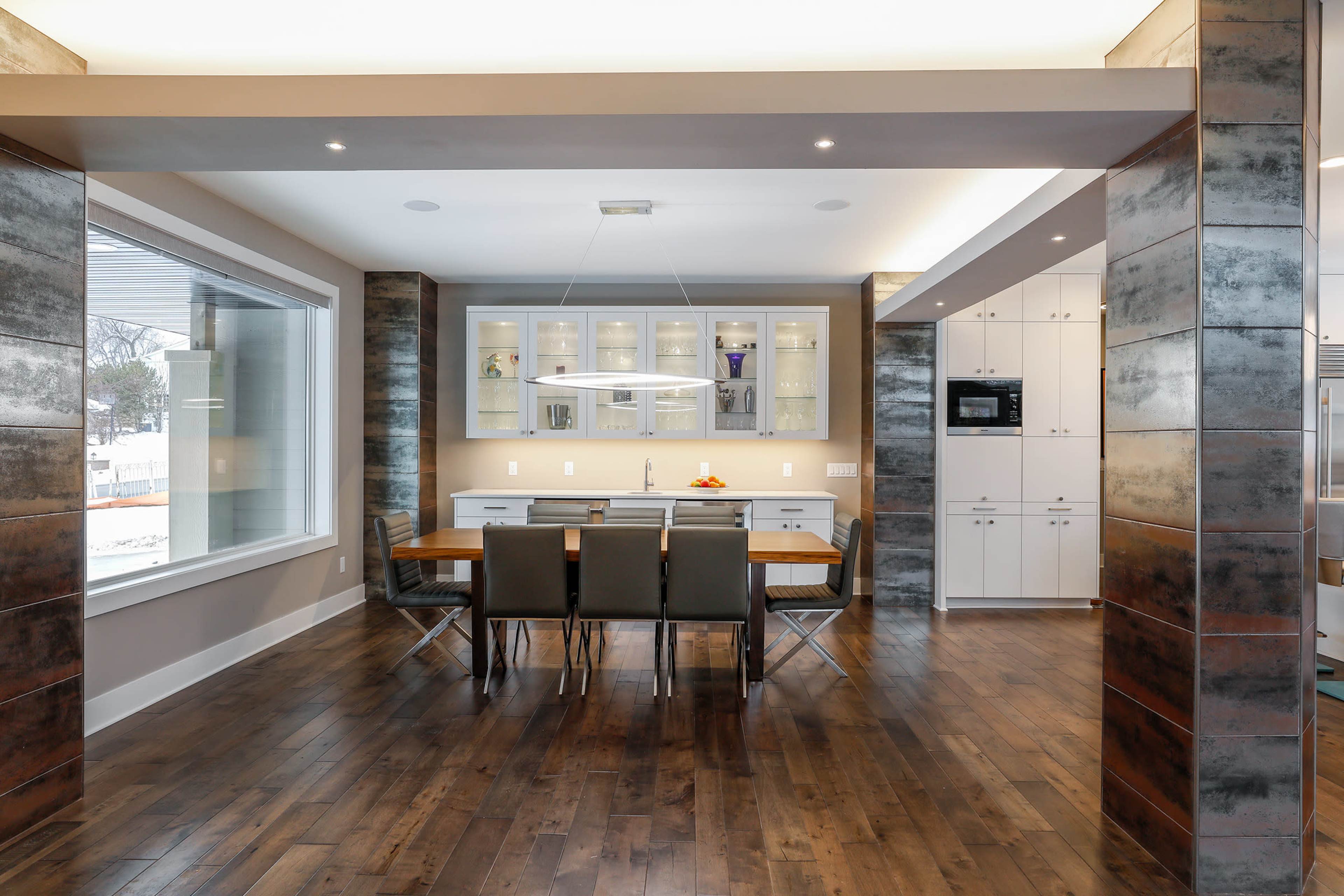 A modern kitchen and dining area features a wooden table with chairs, a sleek countertop, and large windows that provide natural light.