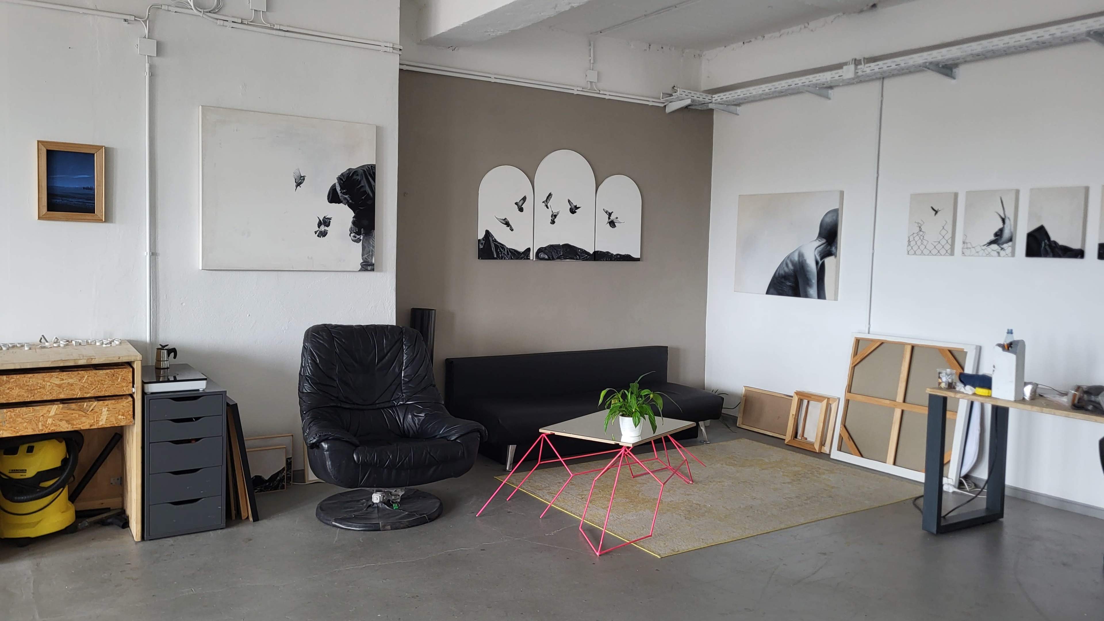 The image shows a modern art studio with a black leather chair, a pink table, and several artworks displayed on the walls.