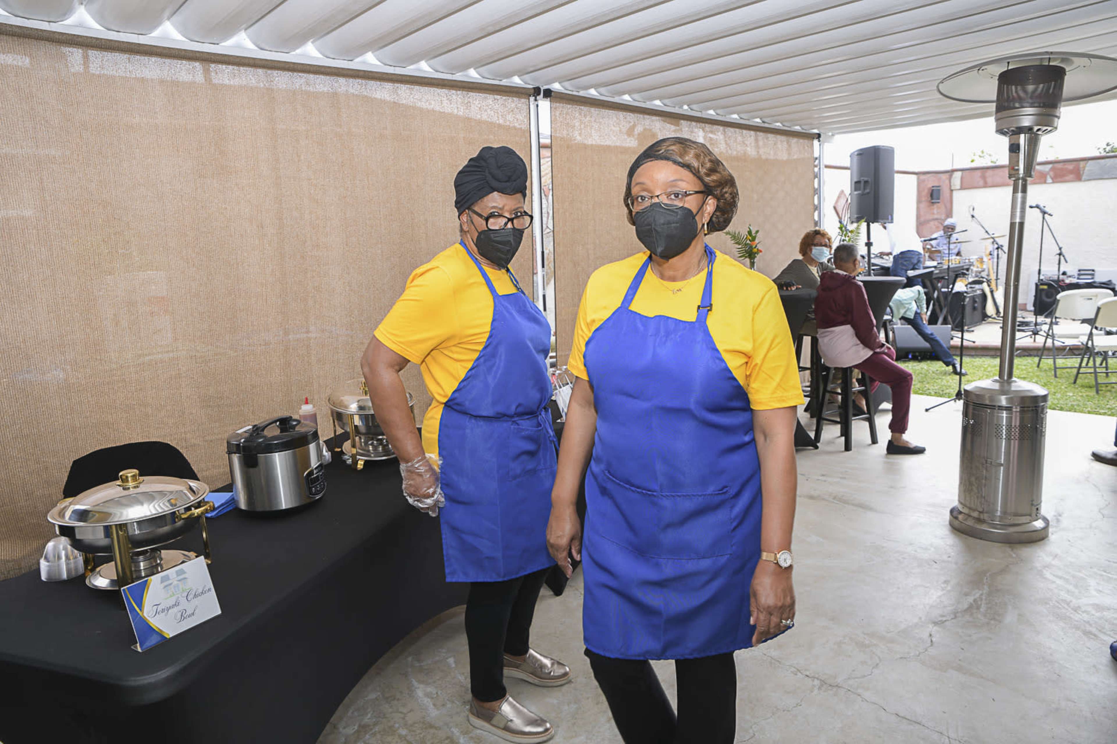 Two women wearing yellow shirts and blue aprons stand behind a table with food containers in an outdoor setting.