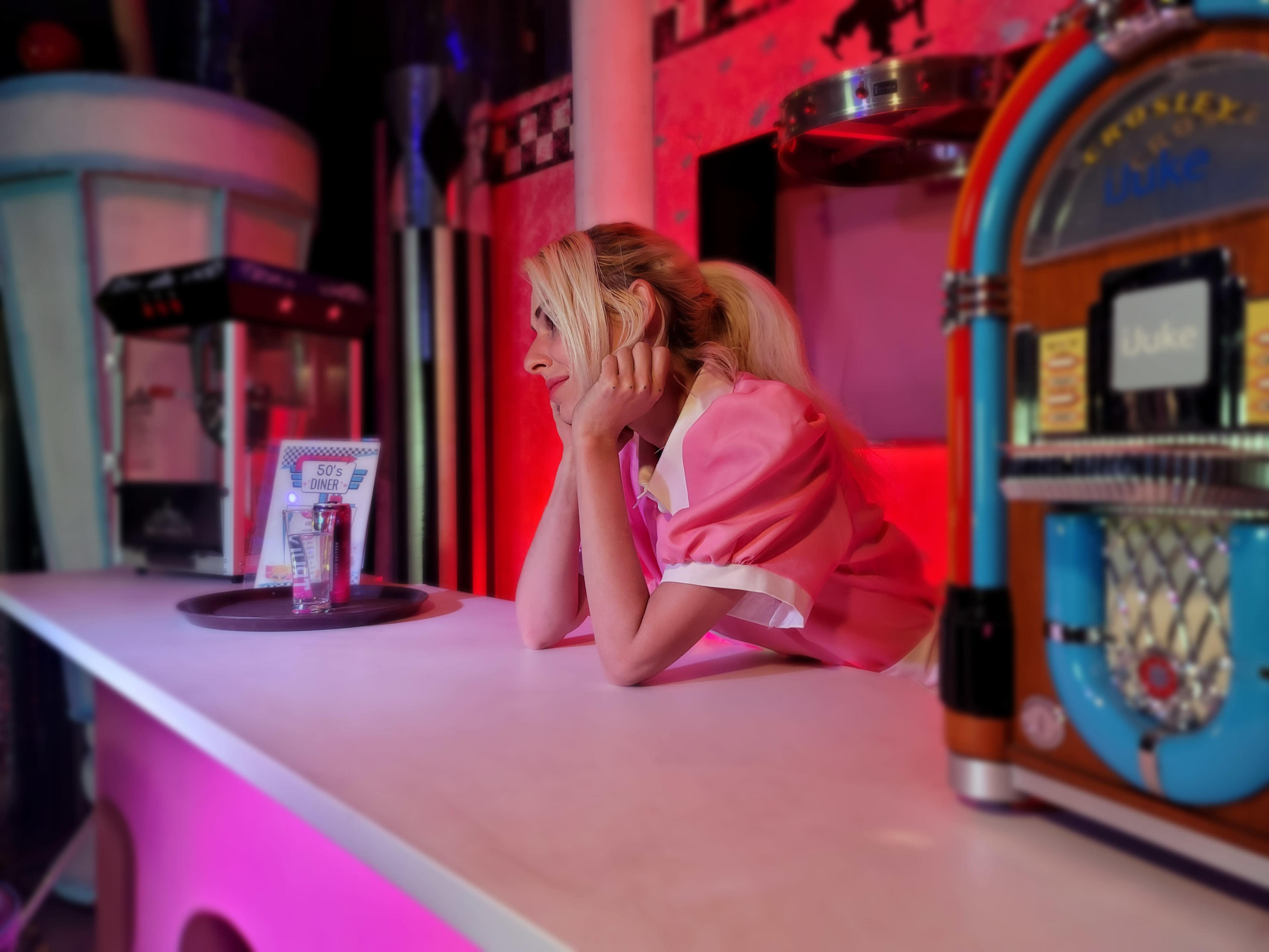 A woman in a pink uniform leans on a countertop near a jukebox in a retro diner setting.