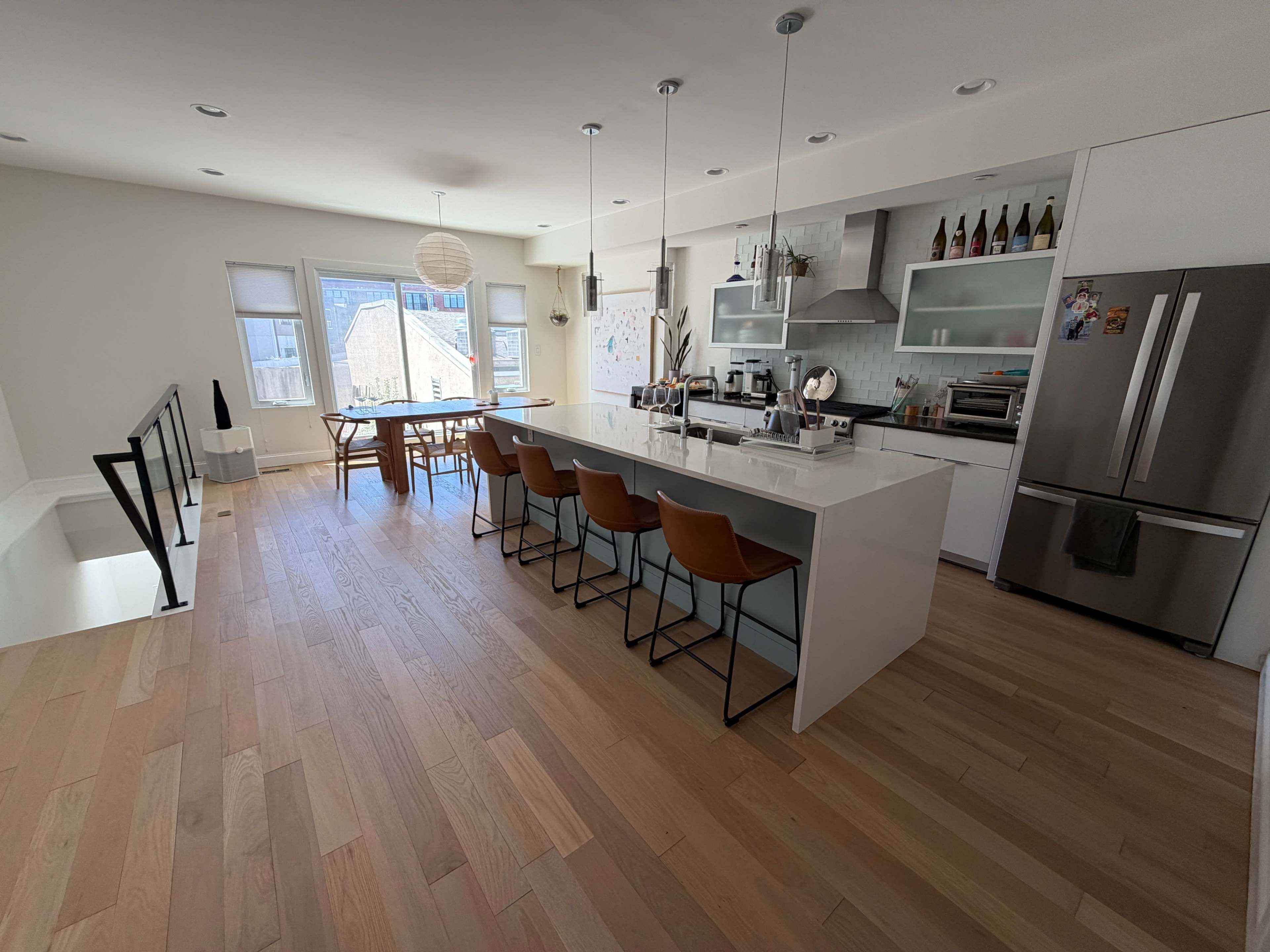 The image shows a modern kitchen with a large island, several bar stools, stainless steel appliances, and abundant natural light from large windows.