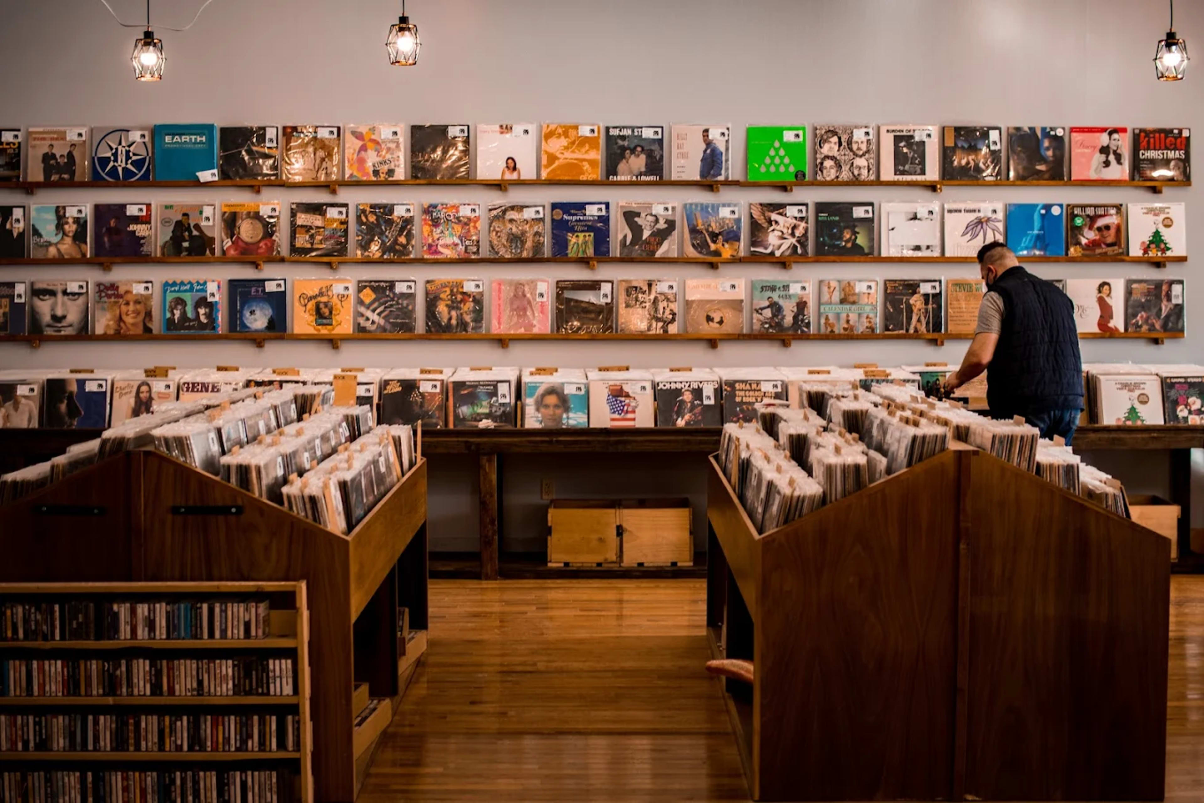 A man is browsing through vinyl records in a music store, which features wooden display tables and a wall lined with album covers.
