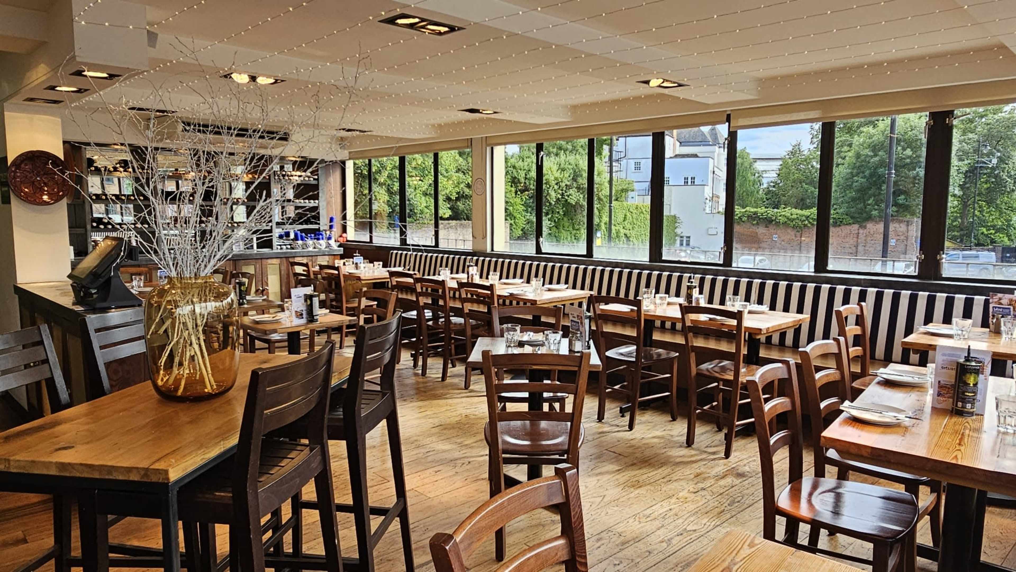 The image shows a spacious restaurant with wooden tables and chairs, large windows, and a bar area lined with bottles.
