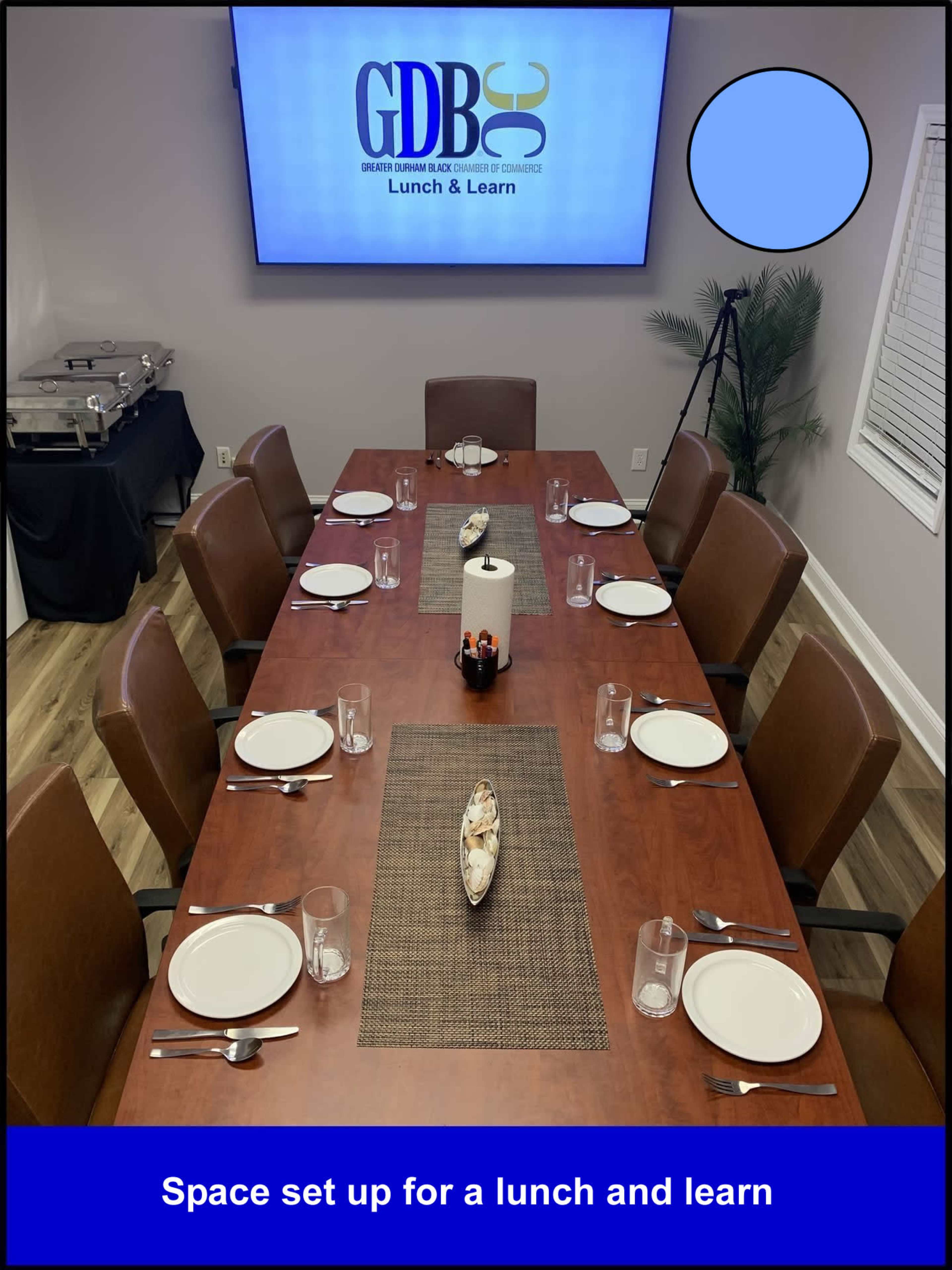 A conference room table is set for a lunch and learn event, with plates, glasses, and a centerpiece, alongside a presentation screen displaying the event title.