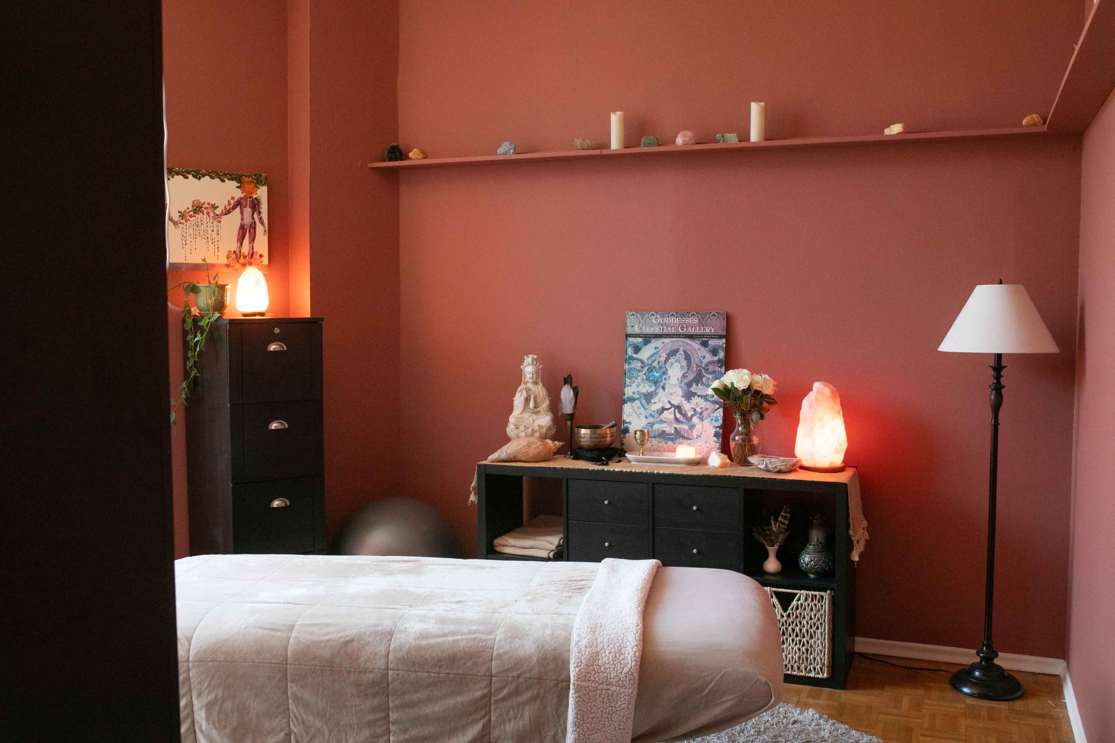 The image shows a serene massage therapy room with a massage table, decorative plants, soft lighting, and various wellness items on display.