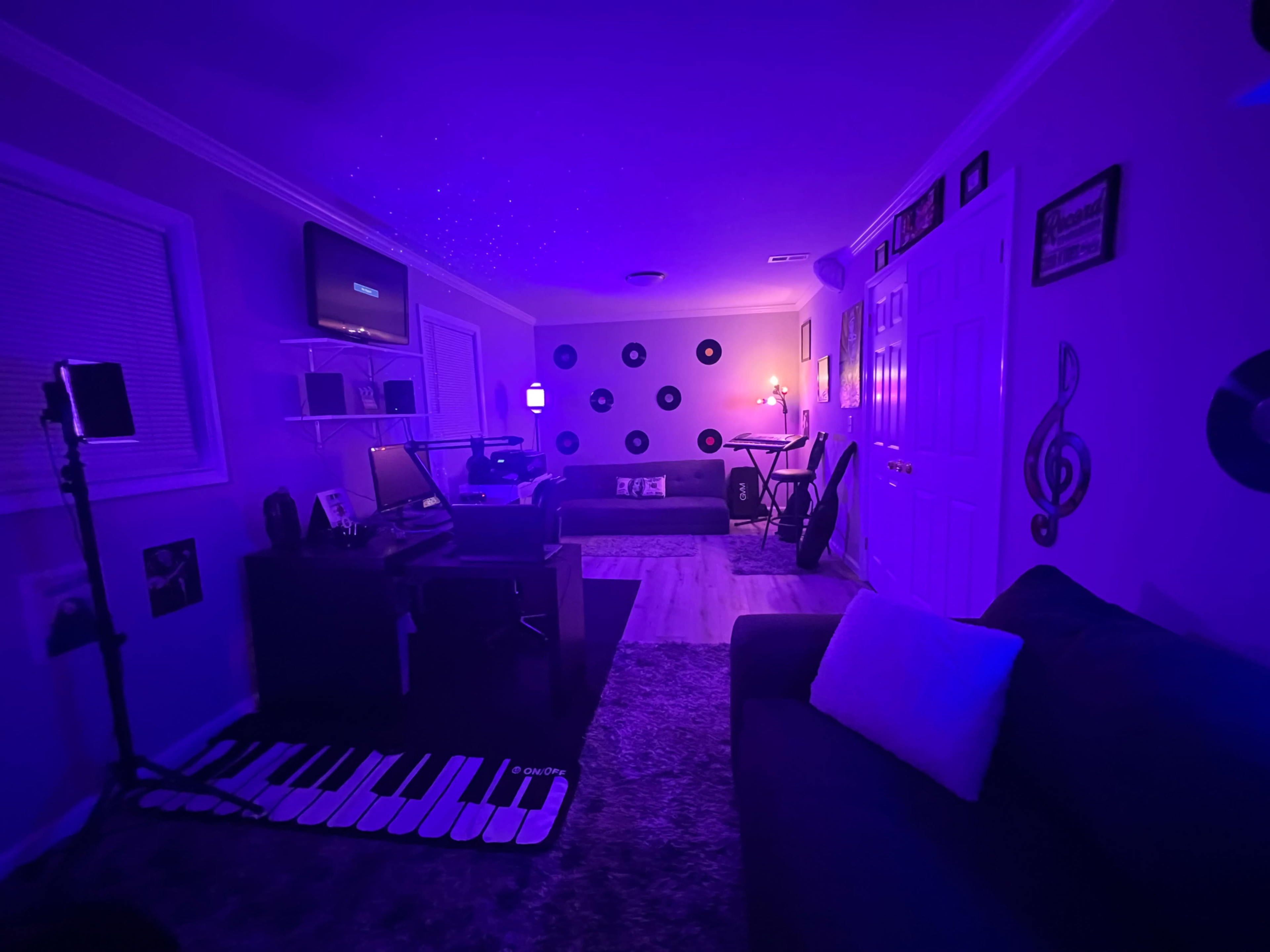 The image shows a dimly lit room with purple lighting, featuring a couch, a keyboard mat, a computer setup, and various music-themed decorations on the walls.