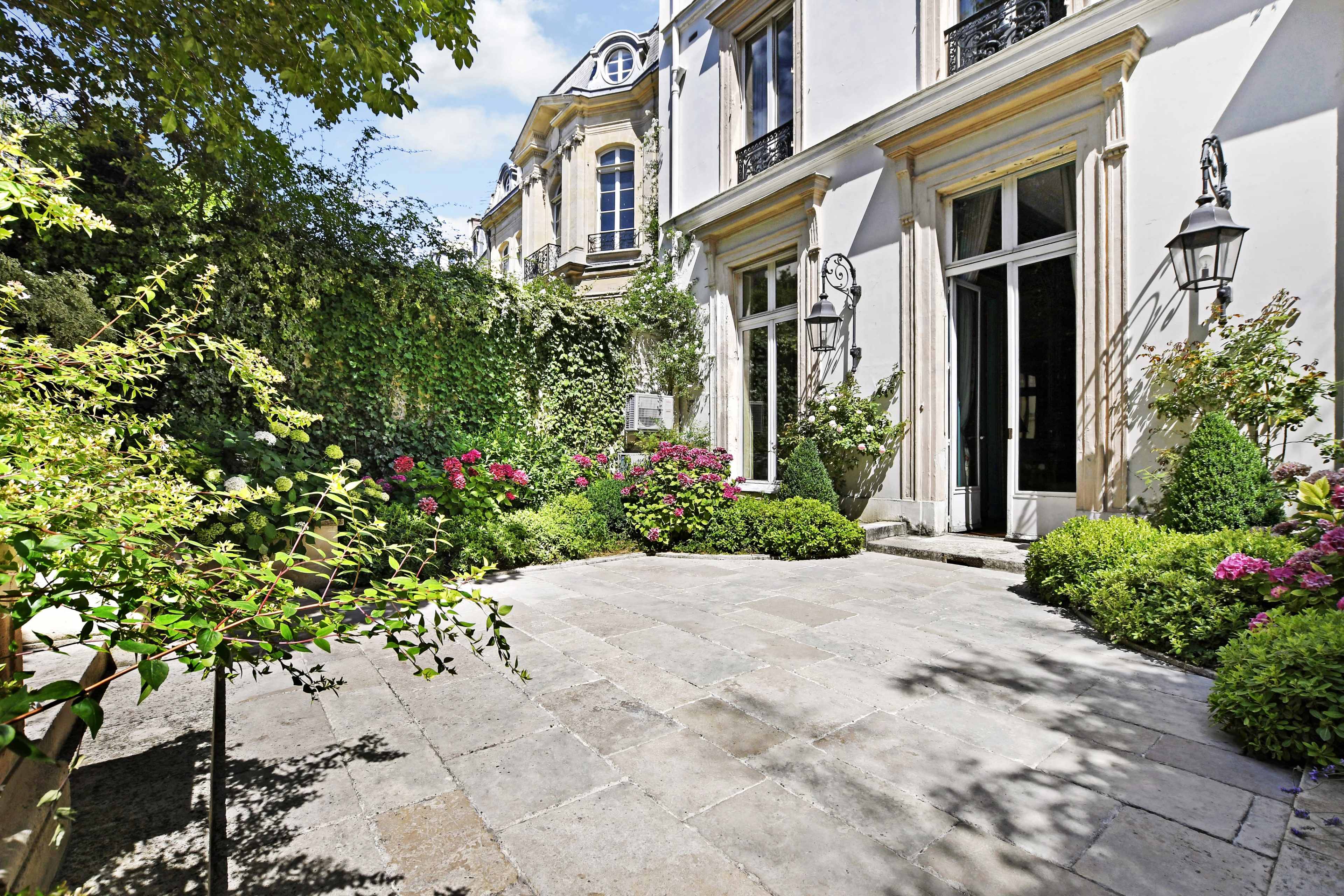 A stone patio surrounded by lush greenery and flowering plants leads to a grand building with large French doors.